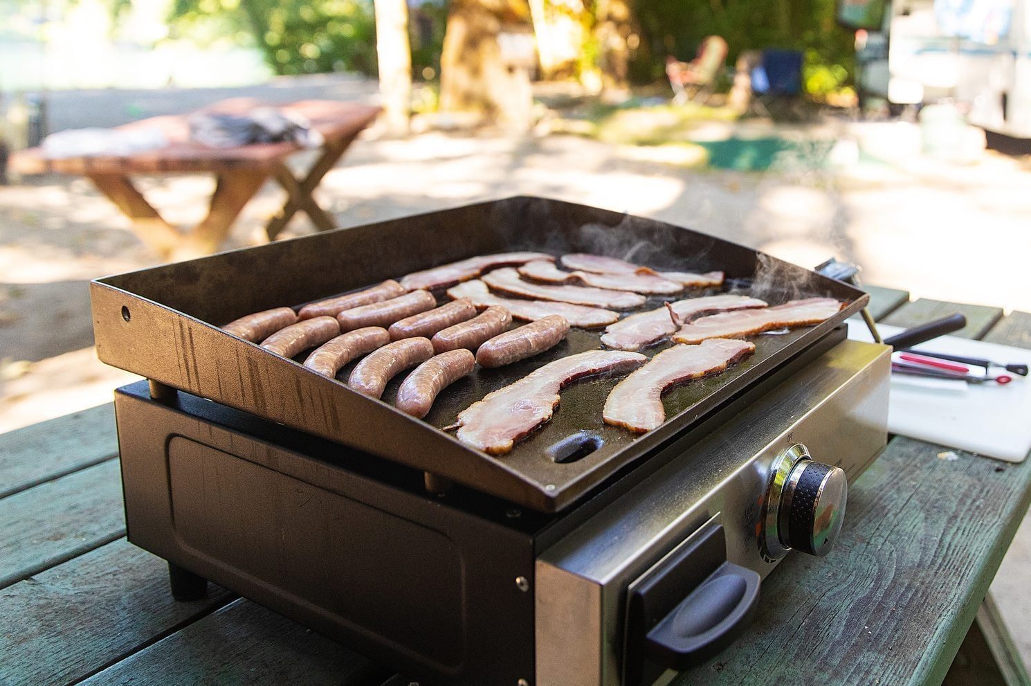 A griddle with cooking sausages and bacon emits steam, positioned on a wooden table in a sunny outdoor setting with nature in the background