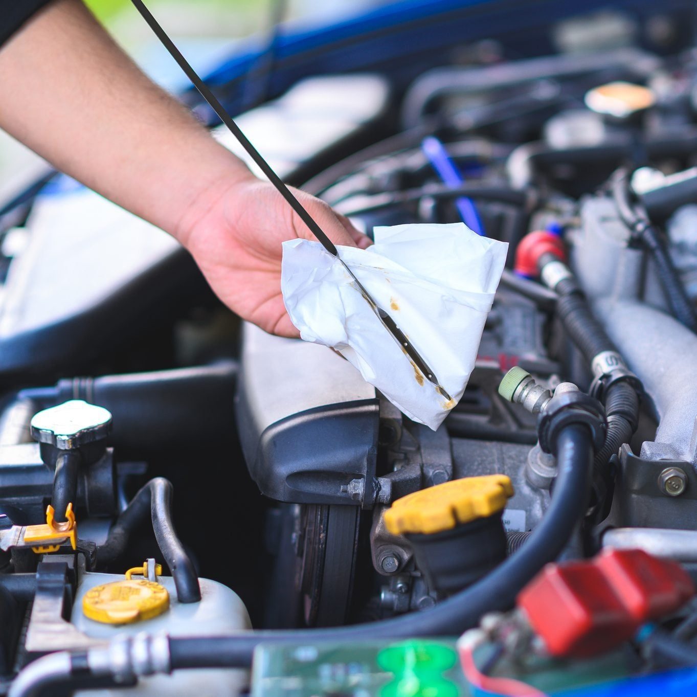 A hand holds an oil dipstick covered with a white tissue, inspecting engine oil levels under a car's hood surrounded by various engine compo