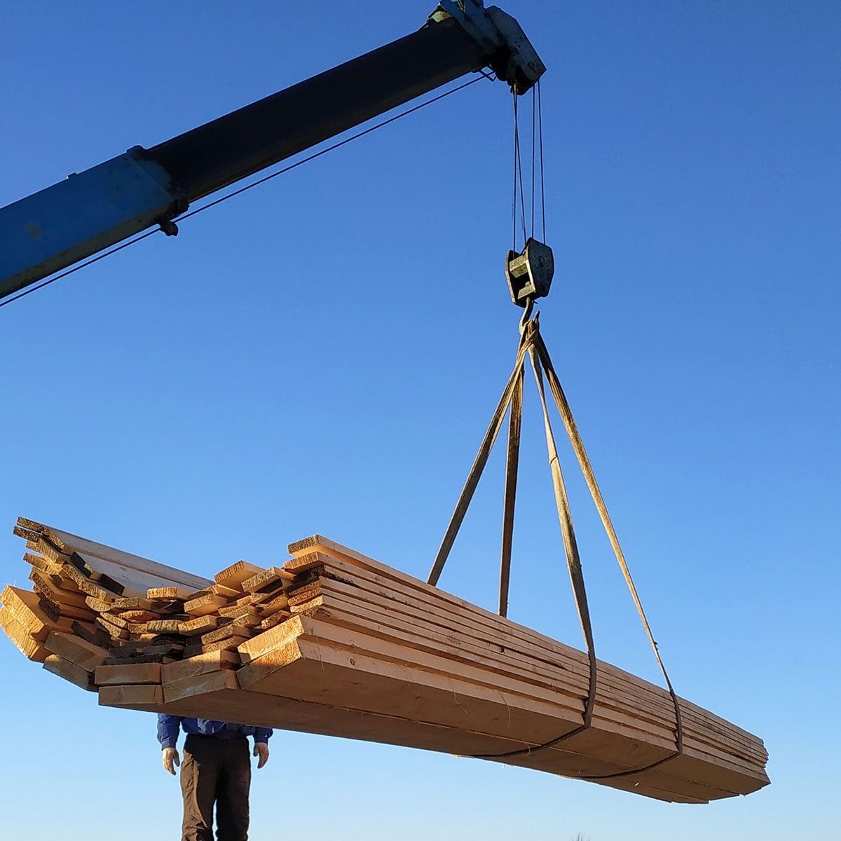 A crane lifts a large bundle of wooden planks, suspended by straps, set against a clear blue sky.