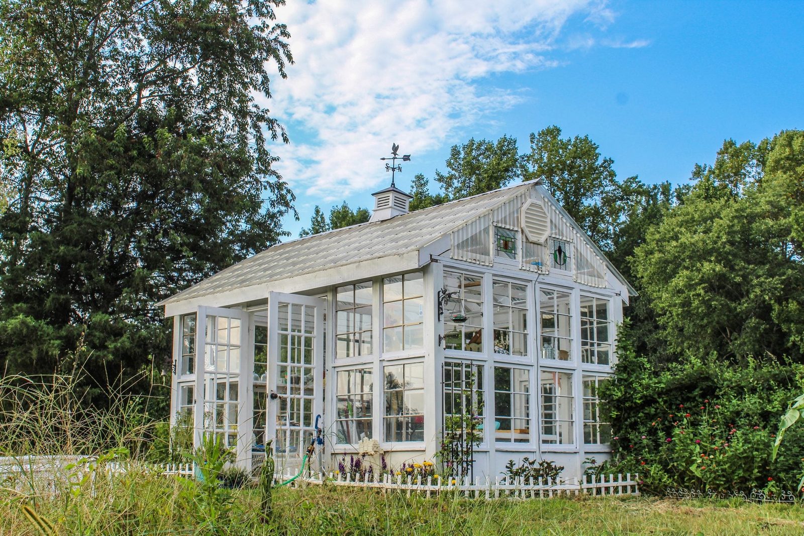 A white greenhouse stands with open doors, showcasing plants inside, surrounded by tall grass and trees under a blue sky with scattered clou