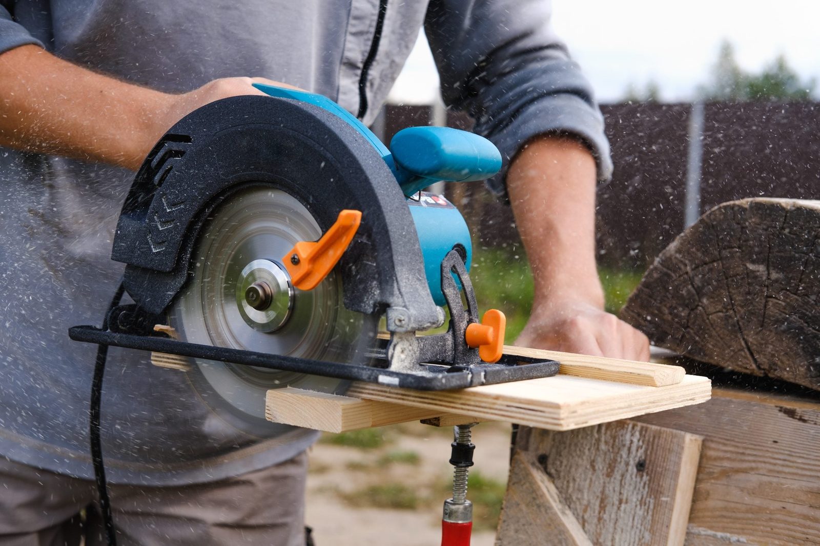 A circular saw cuts through a wooden plank, creating sawdust in an outdoor setting with blurred background elements.