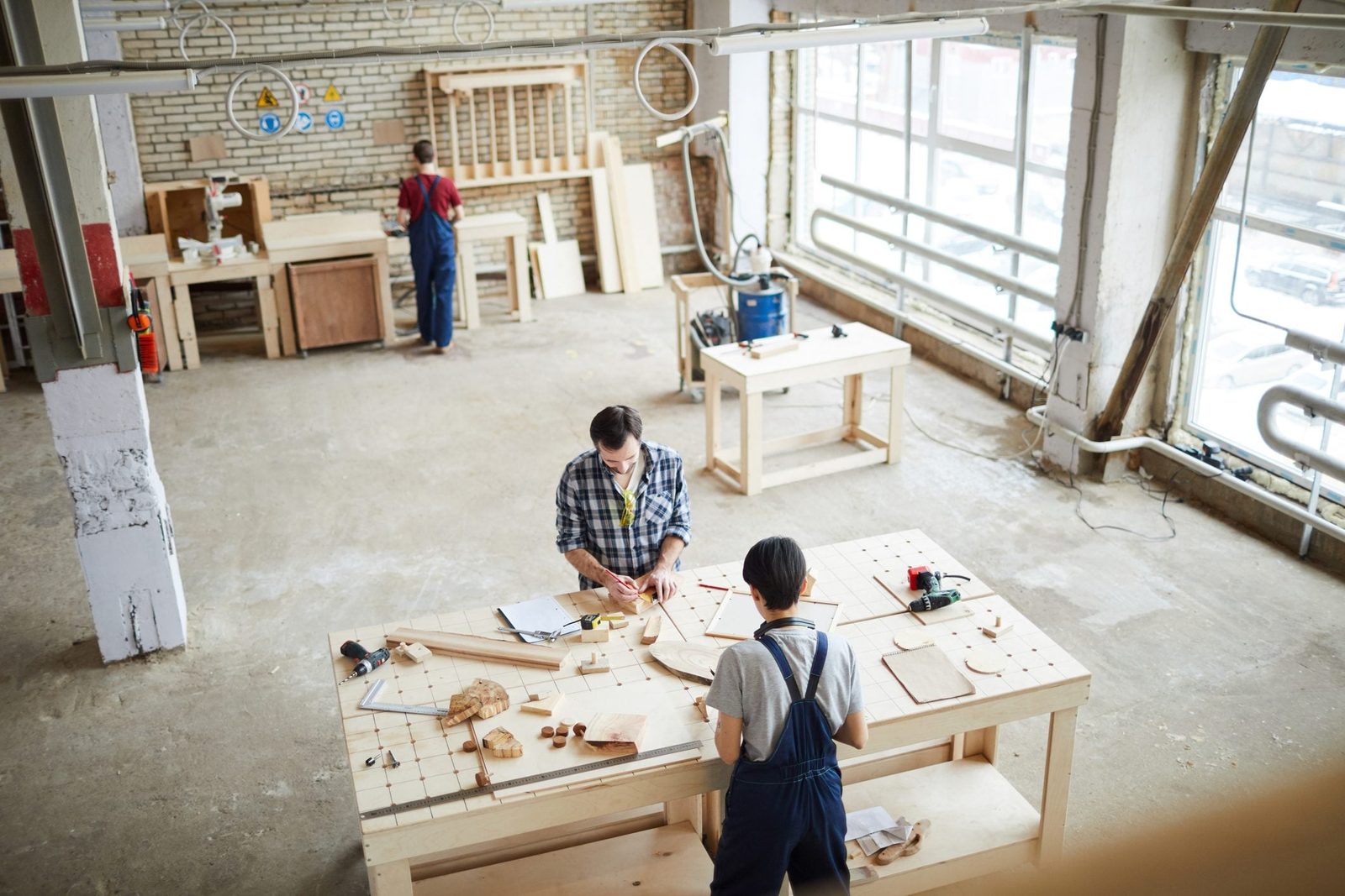A man and a child work on woodworking projects at a large table in a well-lit workshop, surrounded by tools and wooden materials.