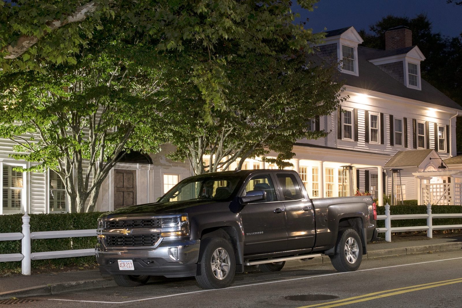 A black pickup truck is parked on a street, illuminated by warm outdoor lights, beside a white house surrounded by trees at night.