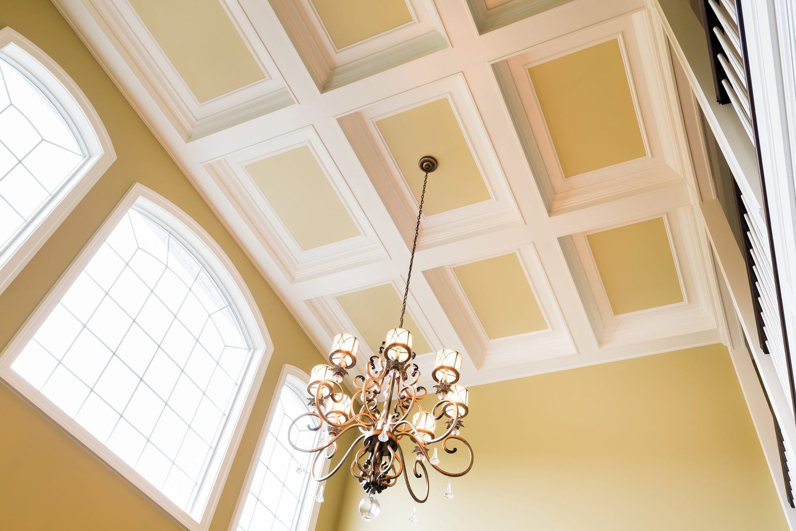 A chandelier hangs from a coffered ceiling, illuminating the spacious room with large arched windows and soft yellow walls.