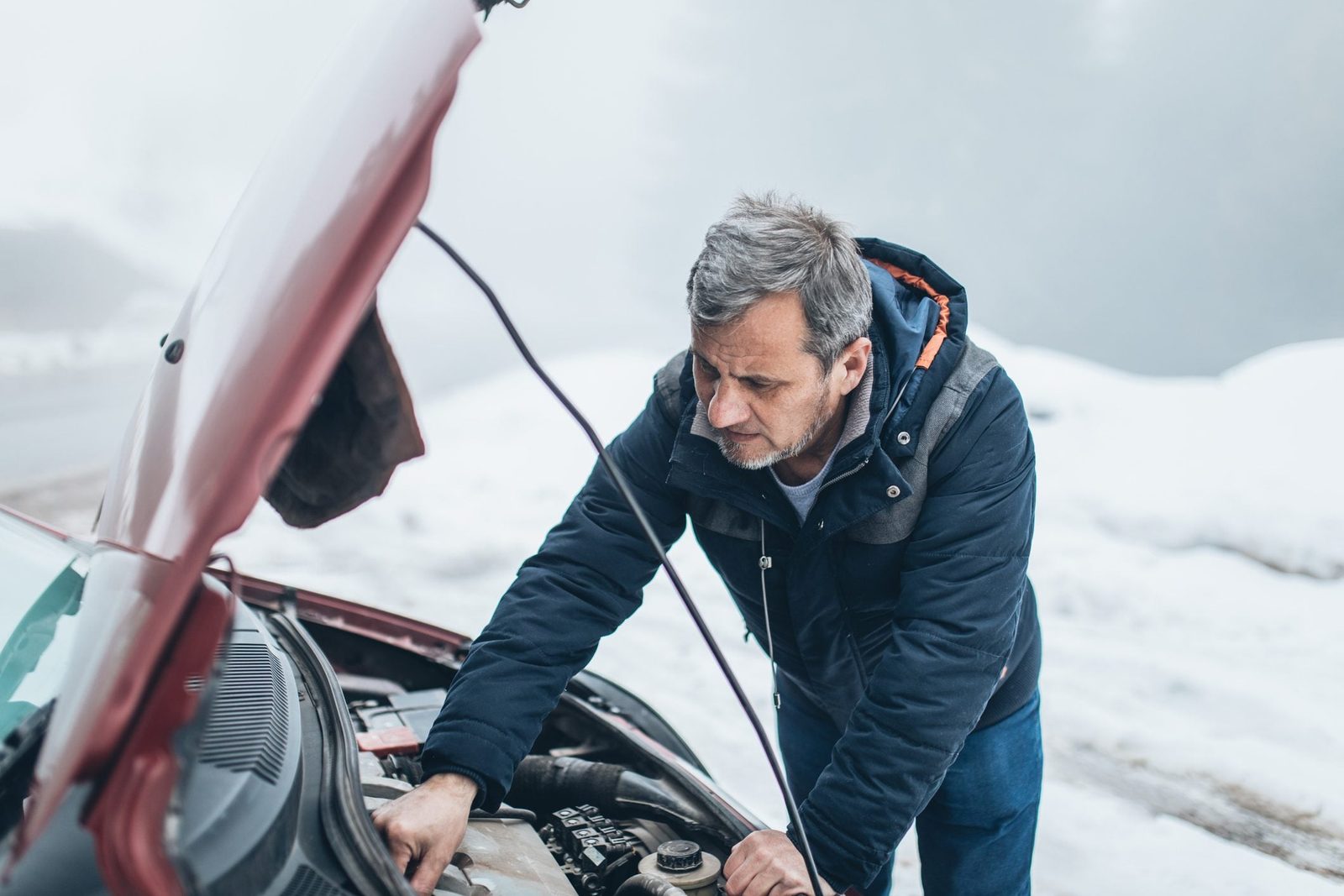 A man inspects a car engine under the hood while standing in a snowy, foggy environment.