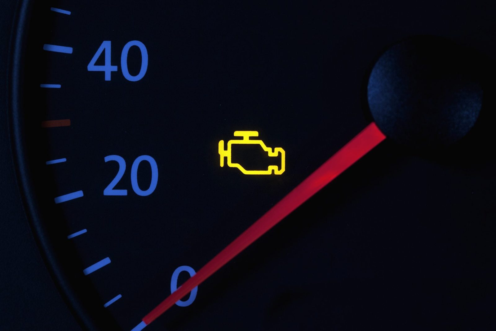 A car dashboard displays a yellow engine warning light alongside a red gauge marker, indicating a potential engine issue in a dark environme