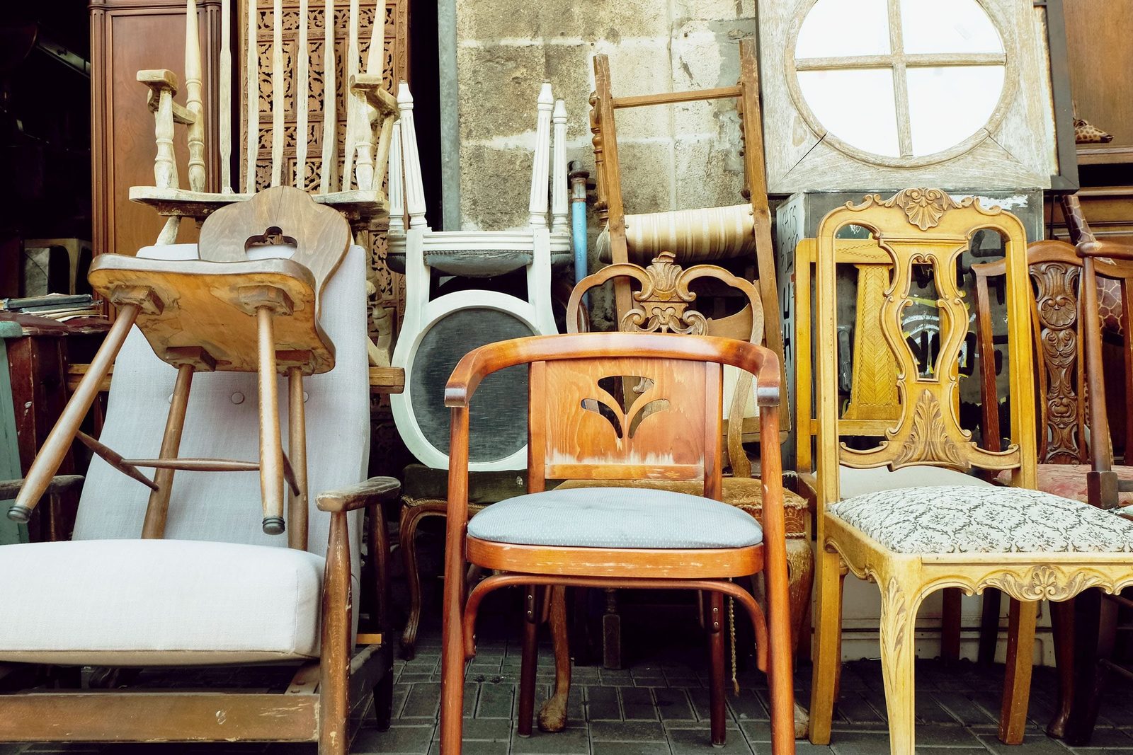 A variety of chairs stands upright, some stacked or leaning, in a dimly lit shop filled with assorted wooden furniture.