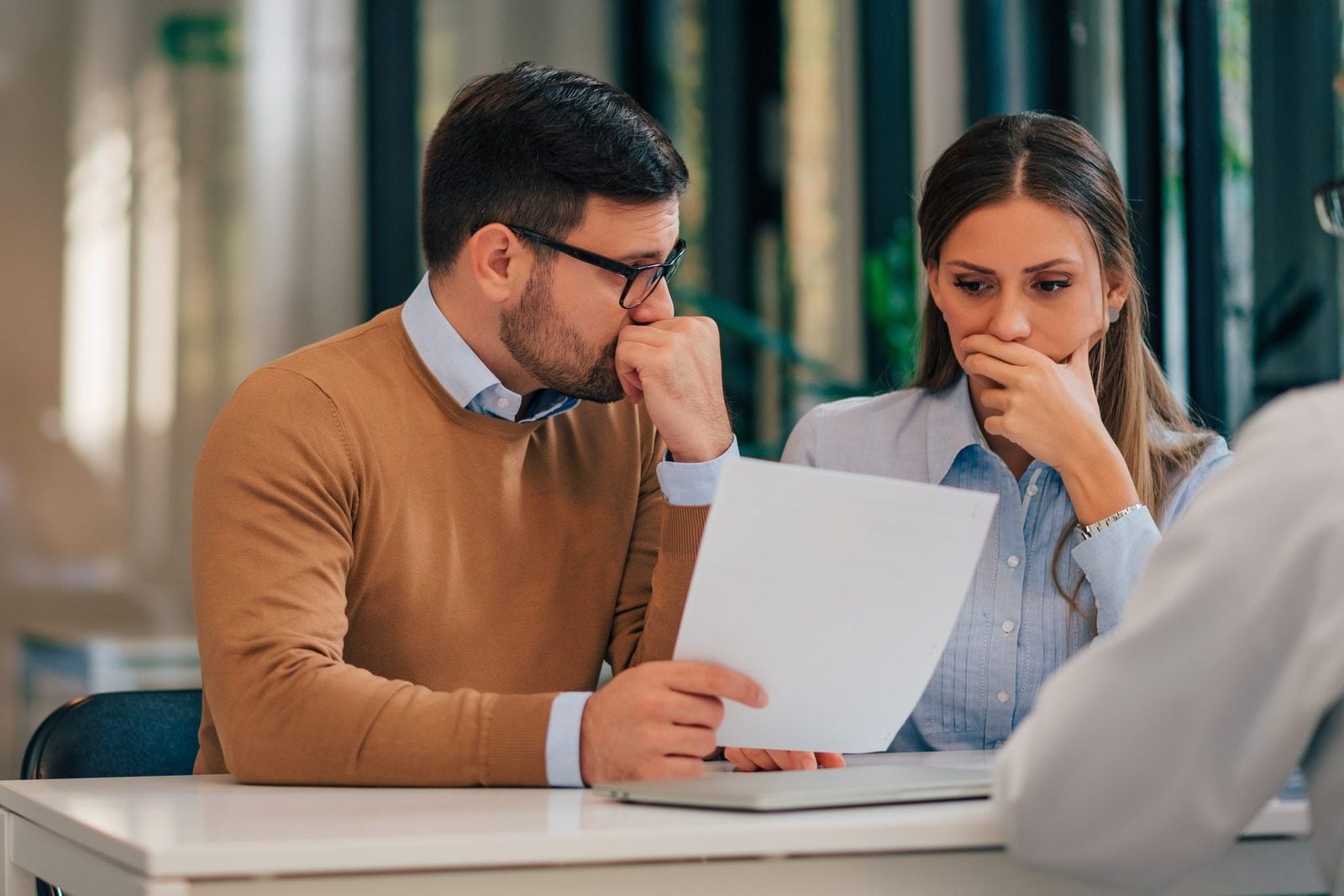A man and woman sit at a table, both concerned while examining a document, with a blurred figure across from them in a bright, modern settin