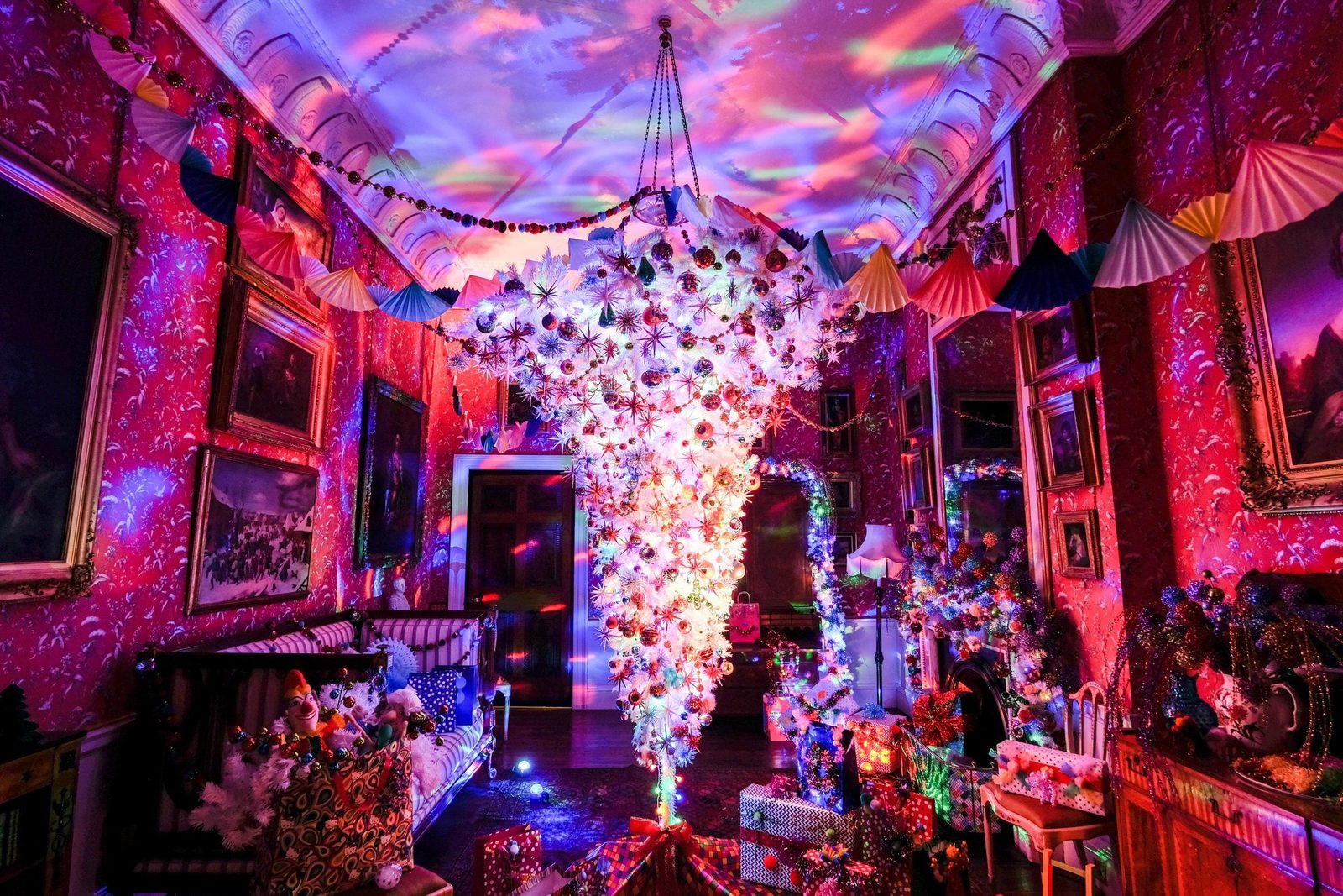 A brightly decorated room features a colorful upside-down Christmas tree and numerous presents, illuminated by vibrant lights reflecting off
