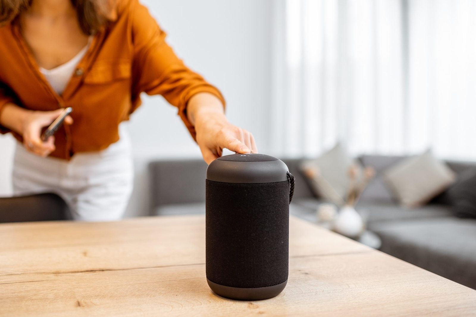 A woman touches a black speaker on a wooden table, while holding a phone. A cozy living room with a gray couch and decorative pillows is in