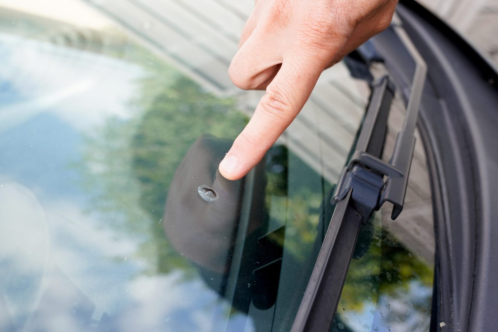 A hand points at a small crack on a car windshield, highlighting potential damage in a reflective, outdoor setting with greenery visible.