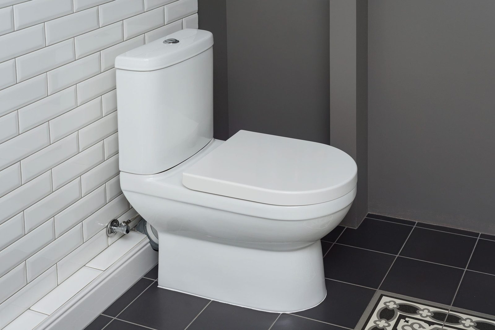 A white toilet sits against a tiled wall in a modern bathroom, featuring a sleek design on dark tile flooring with a decorative mat nearby.