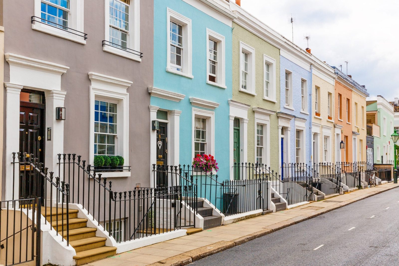 Colorful row houses line a street, featuring flower boxes and gated stairways, set against a cloudy sky and empty pavement.