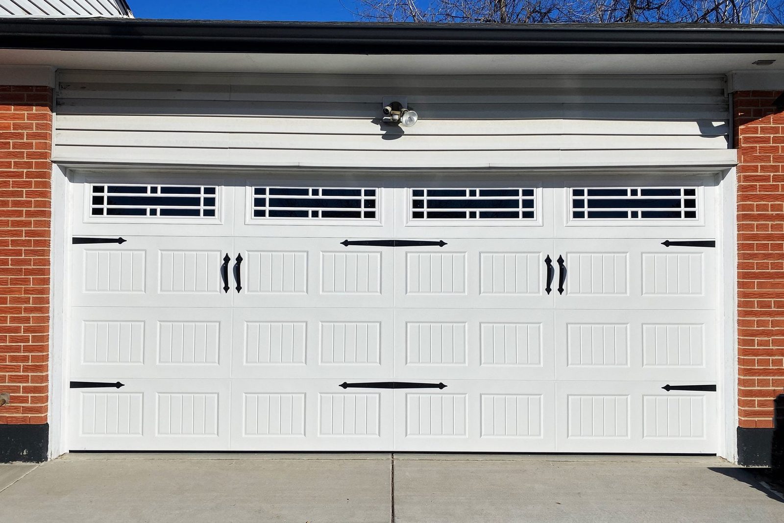 A white garage door with black handles and accents is closed, set against a brick wall and a blue sky, creating a modern suburban scene.