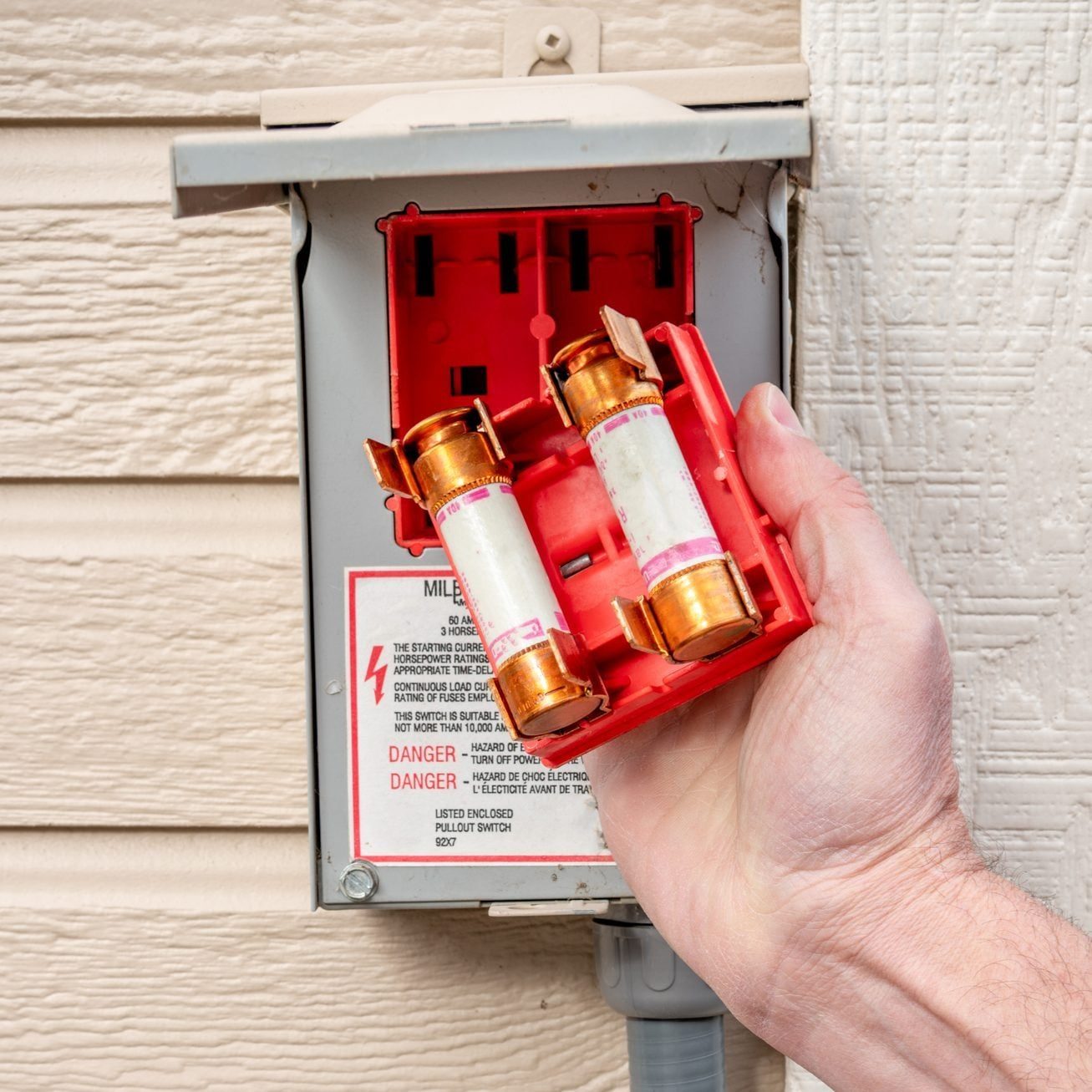A hand holds a red fuse holder with two fuses, positioned near an electrical panel on a textured wall, indicating maintenance activity.