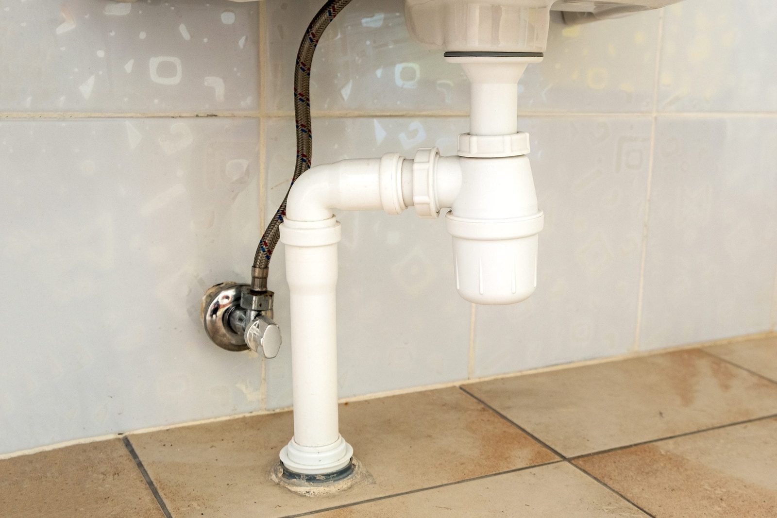 A white plumbing pipe connects to a faucet, draining water into the tiled floor below, beside textured blue wall tiles.