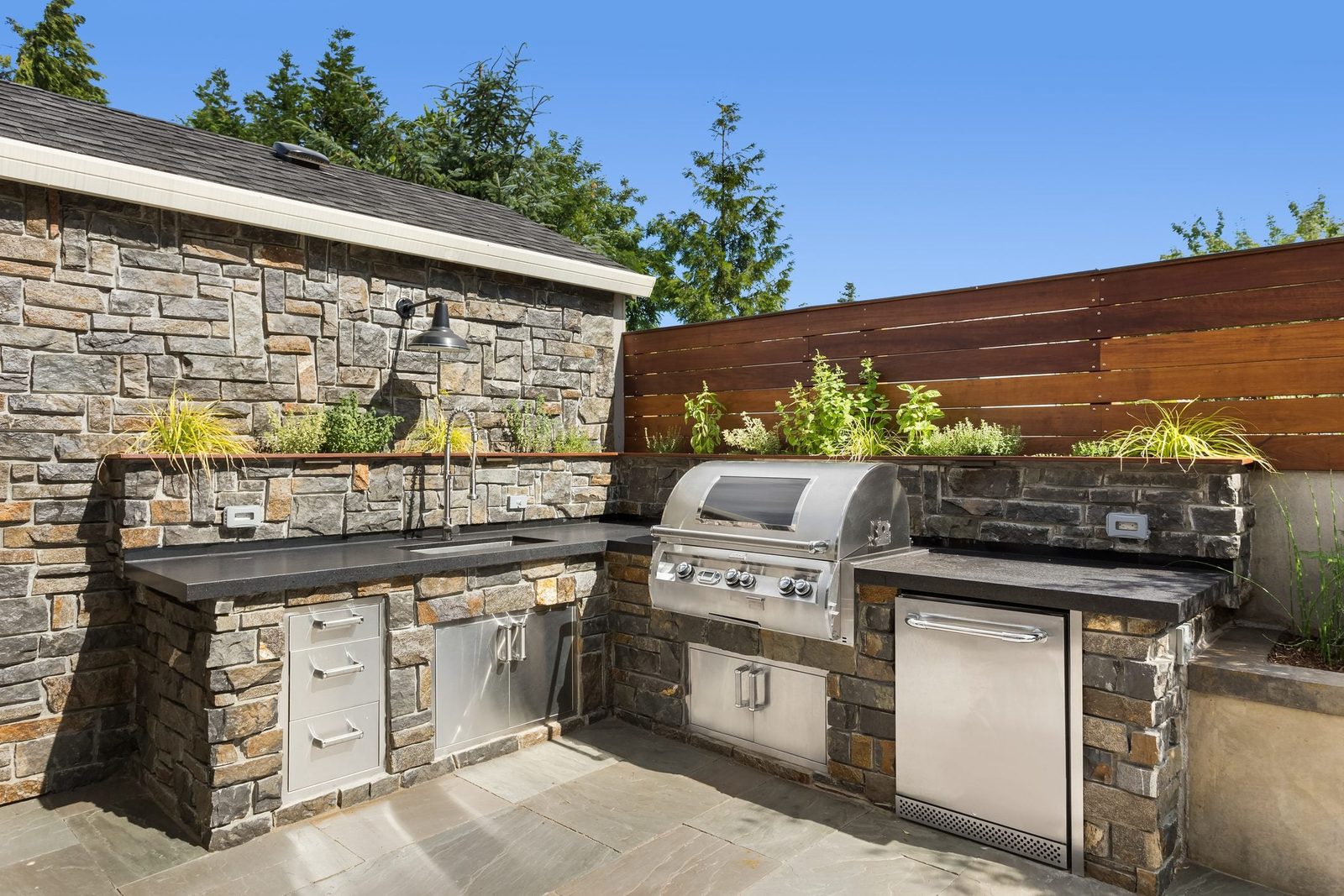 A stone outdoor kitchen features a stainless-steel grill, sink, and storage. Green plants line the back, with a clear blue sky in the backgr