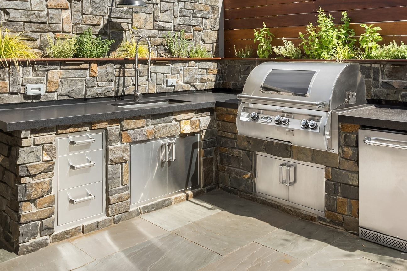 A stainless steel grill sits in a stone outdoor kitchen with a black countertop, surrounded by greenery on a sunny day.