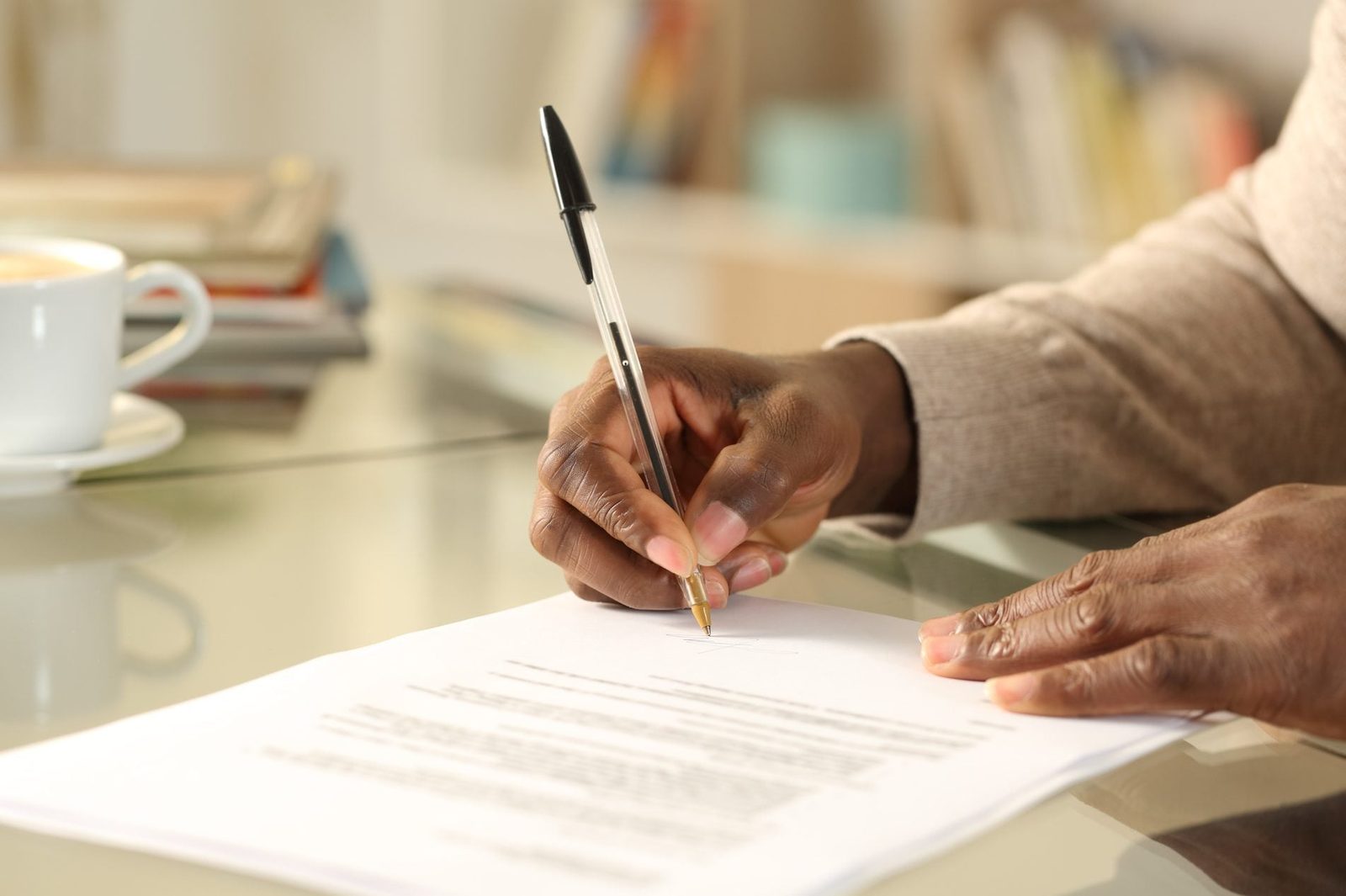 A hand is writing on a document with a pen, while a coffee cup sits nearby on a glass table amid a softly lit background.