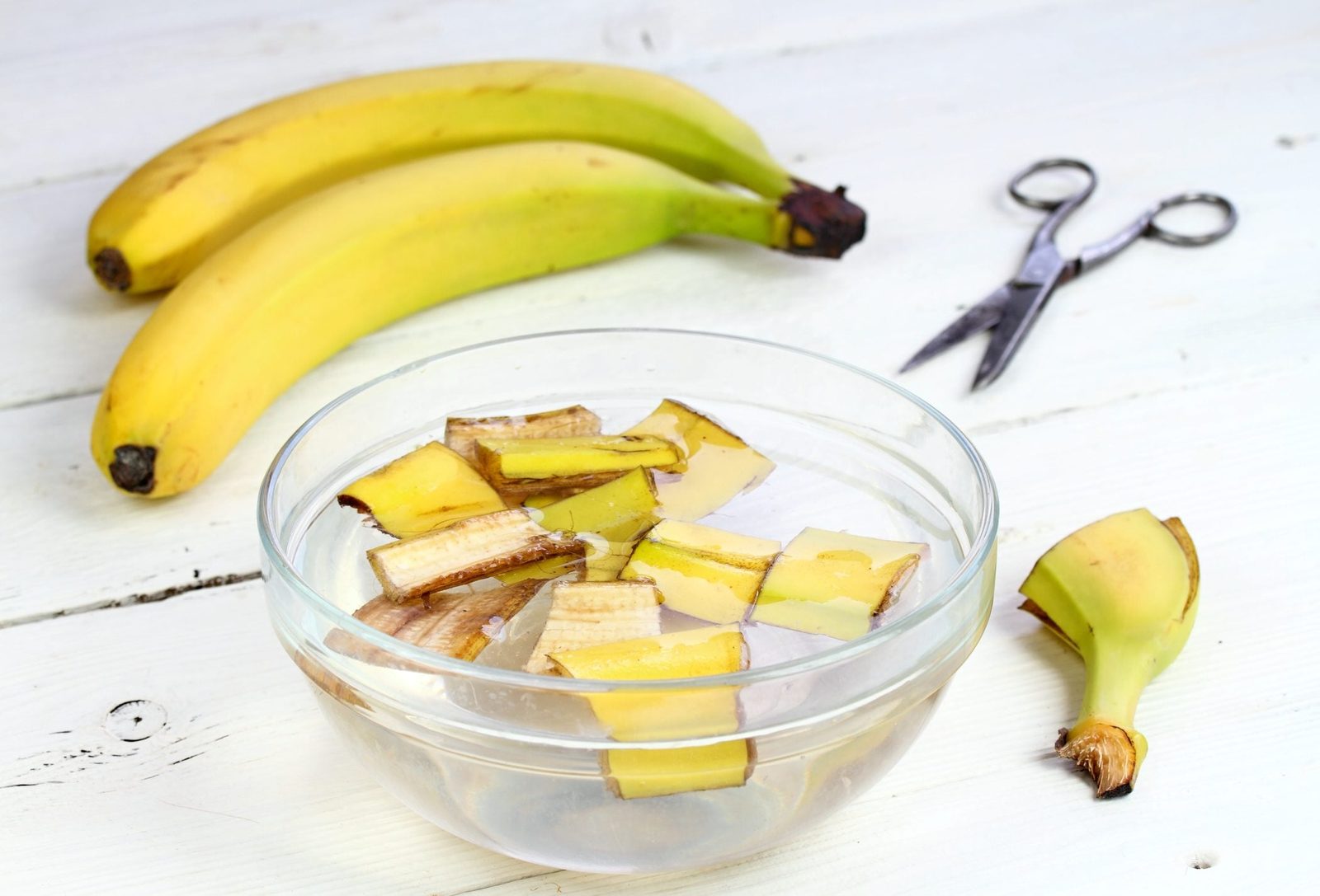 Sliced banana pieces are submerged in water in a clear bowl, placed on a white surface with whole bananas and scissors nearby.