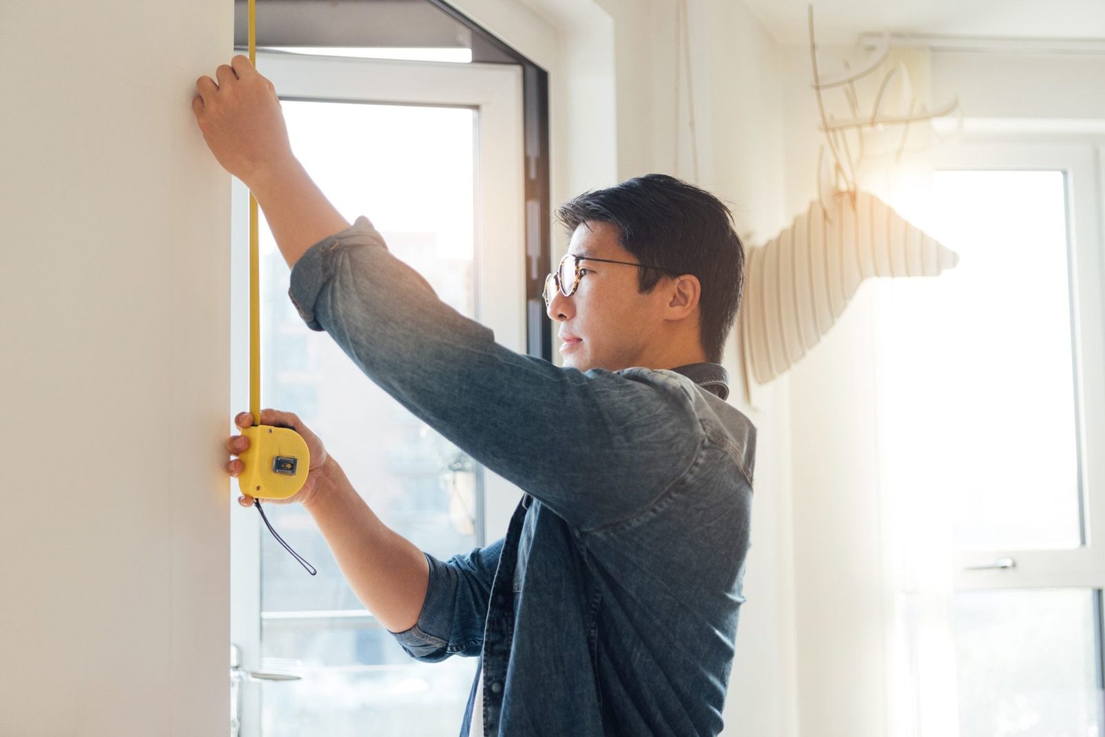 A man holds a tape measure against a wall, measuring height in a well-lit room with a window and a decorative antler-shaped object.