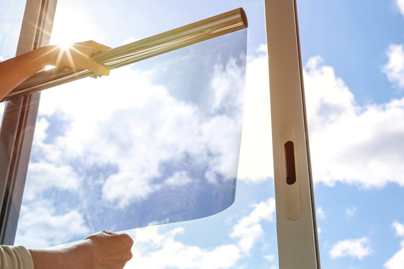 A hand holds a transparent film while applying it to a window, with bright sunlight and a blue sky filled with clouds in the background.