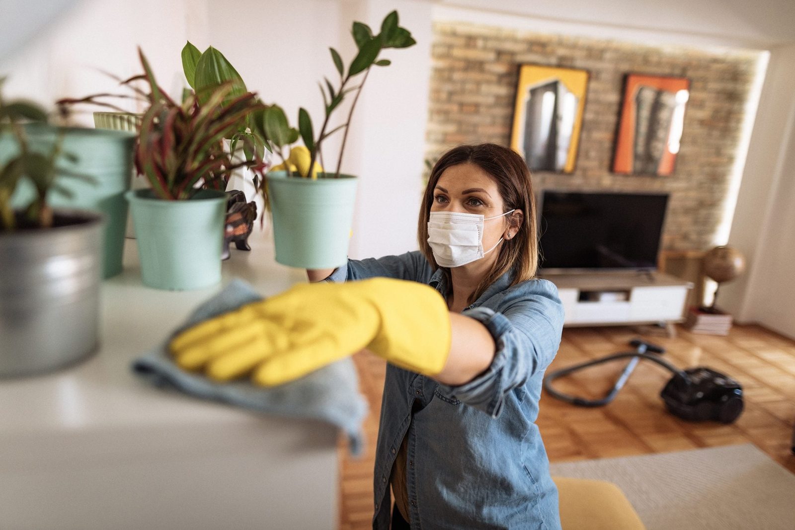 A woman in a mask, wearing yellow gloves, wipes a surface while planting potted plants in a tidy, light-filled living room. A vacuum cleaner