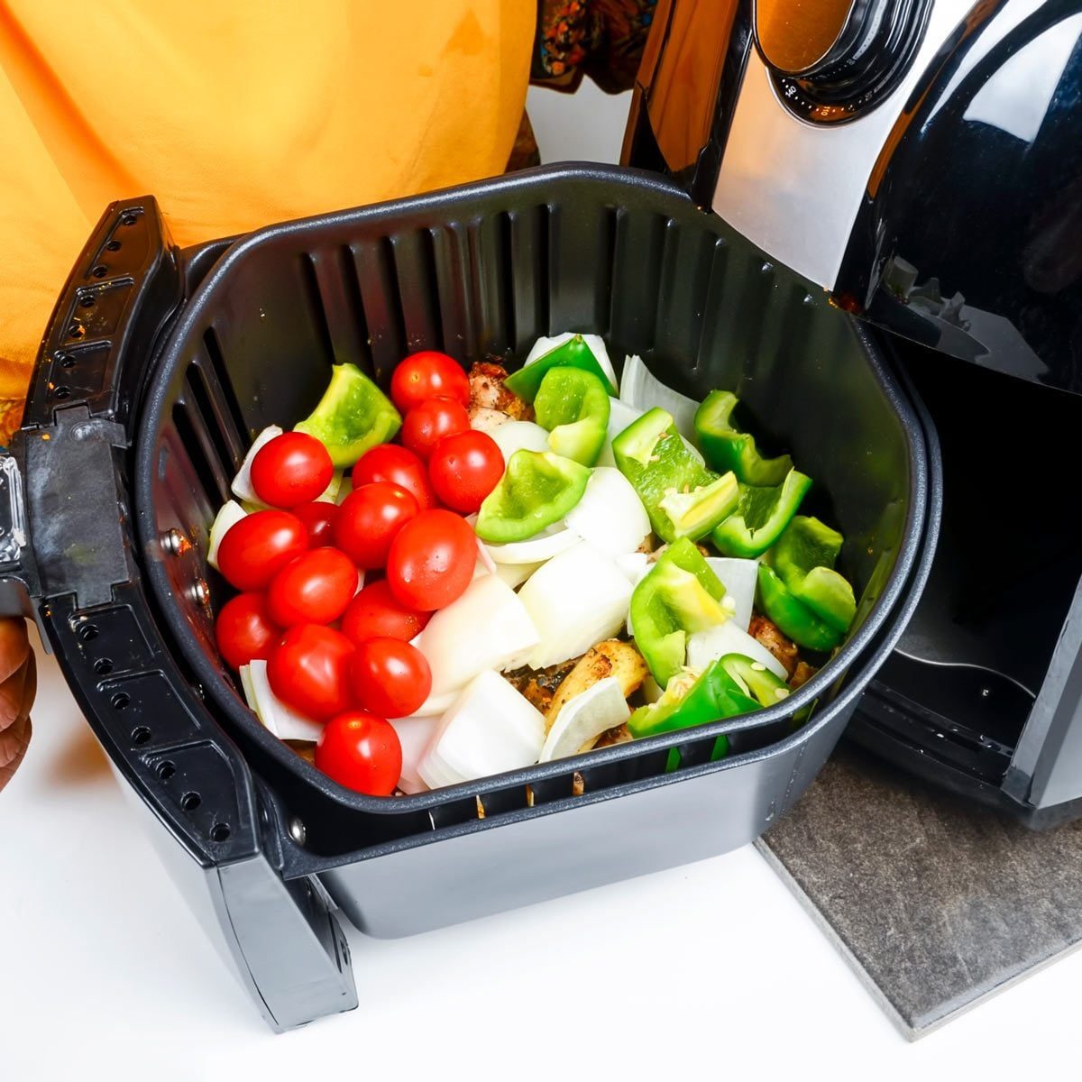 A black air fryer basket contains chopped onions, green bell peppers, and red tomatoes, while a person in an orange shirt holds it next to t