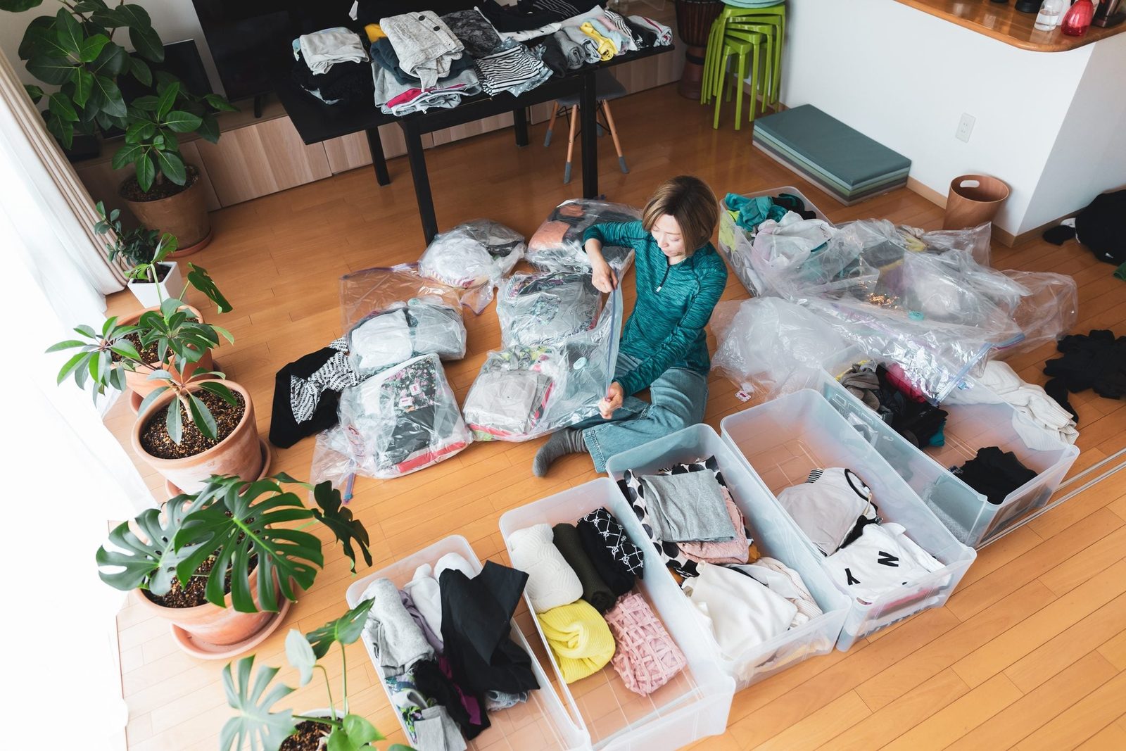 A woman organizes clothes into containers, sorting items on the floor. Potted plants and a table with neatly folded clothes surround her in