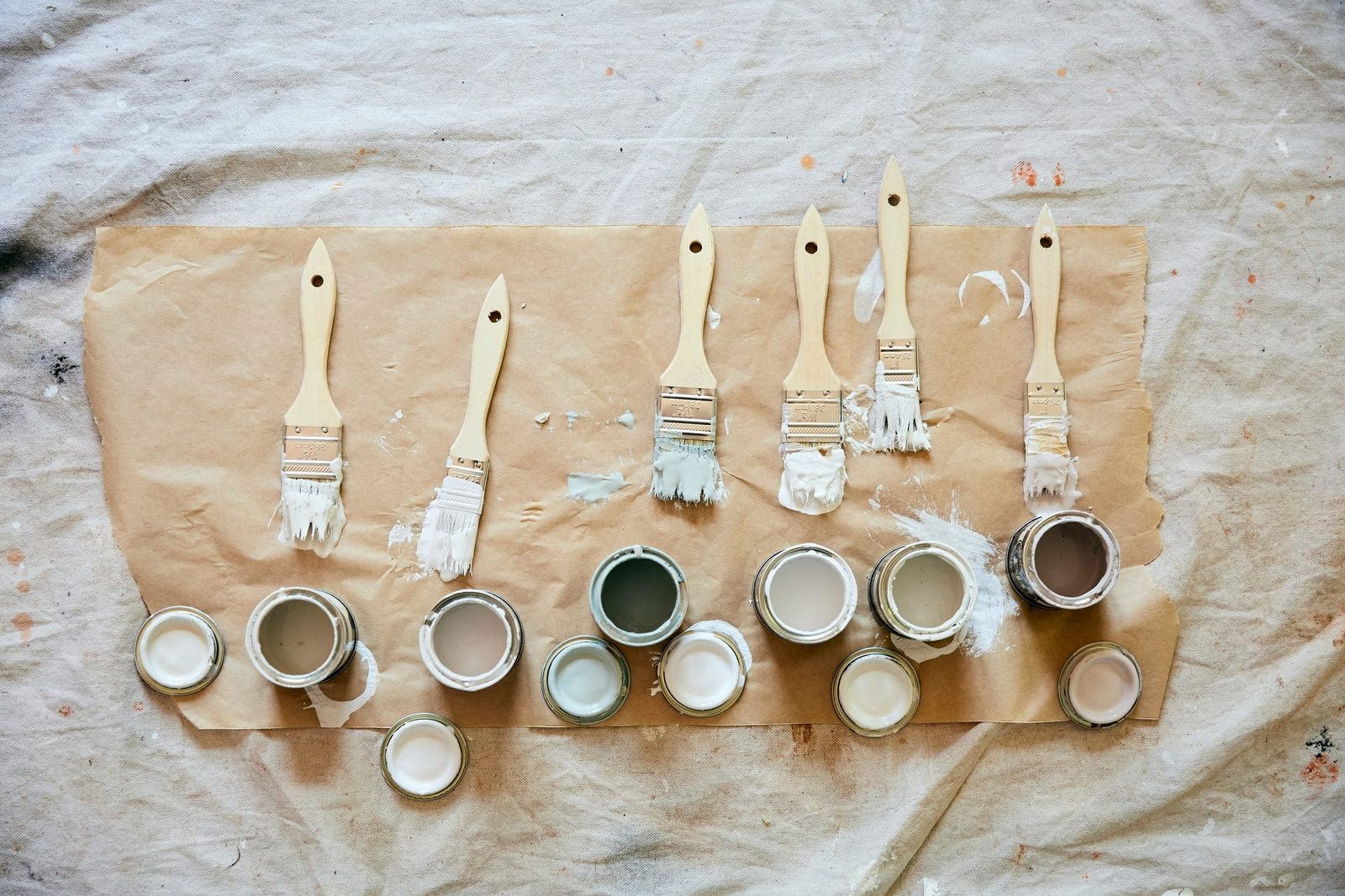 Brushes and paint cans are arranged on a brown paper surface, with paint splatters evident on the surrounding fabric backdrop.