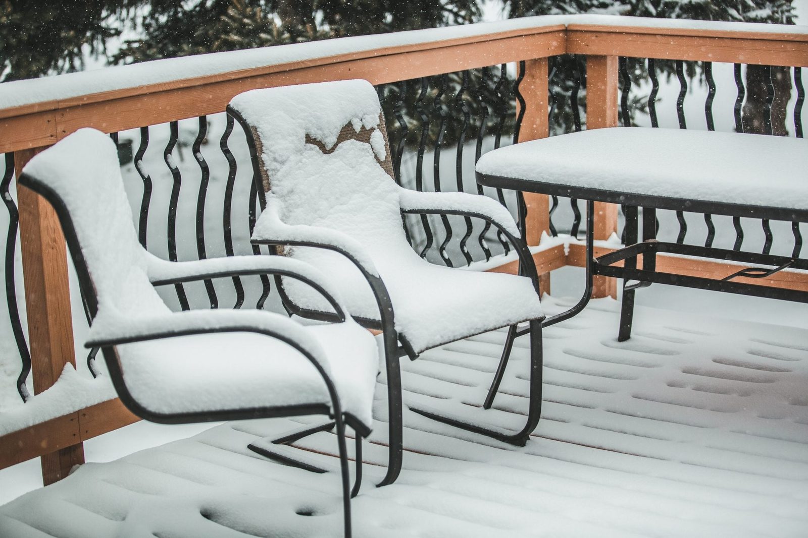 Two snow-covered metal chairs and a table sit on a wooden deck, surrounded by falling snowflakes and a snowy landscape in the background.