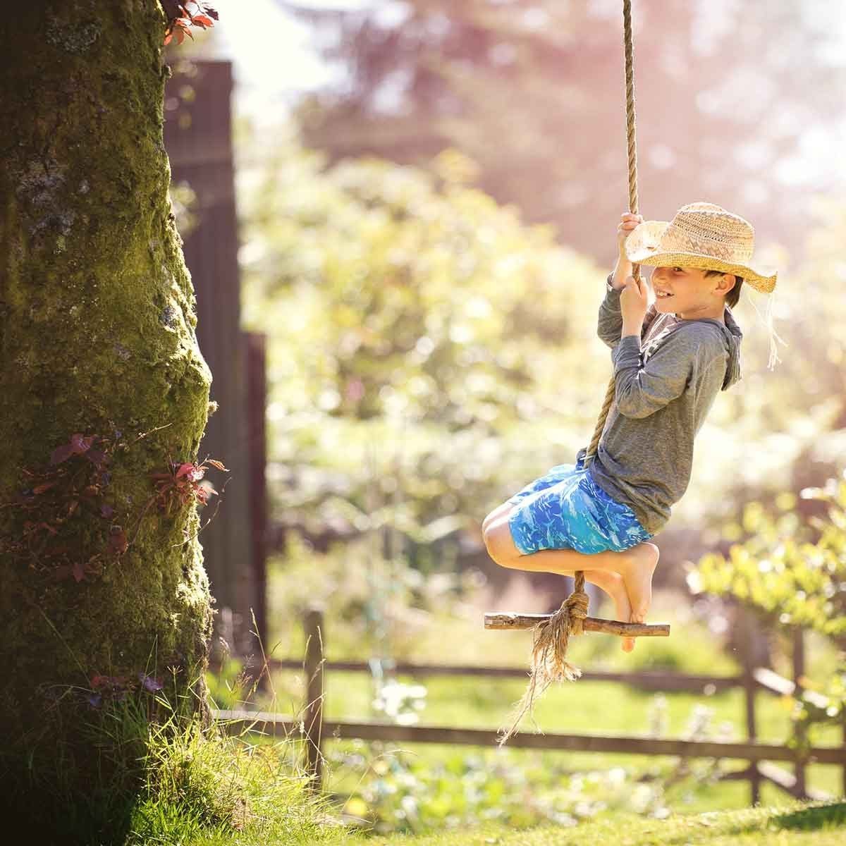 A child swings joyfully on a rope attached to a tree, wearing a straw hat and playful clothing, surrounded by a sunny, green outdoor landsca