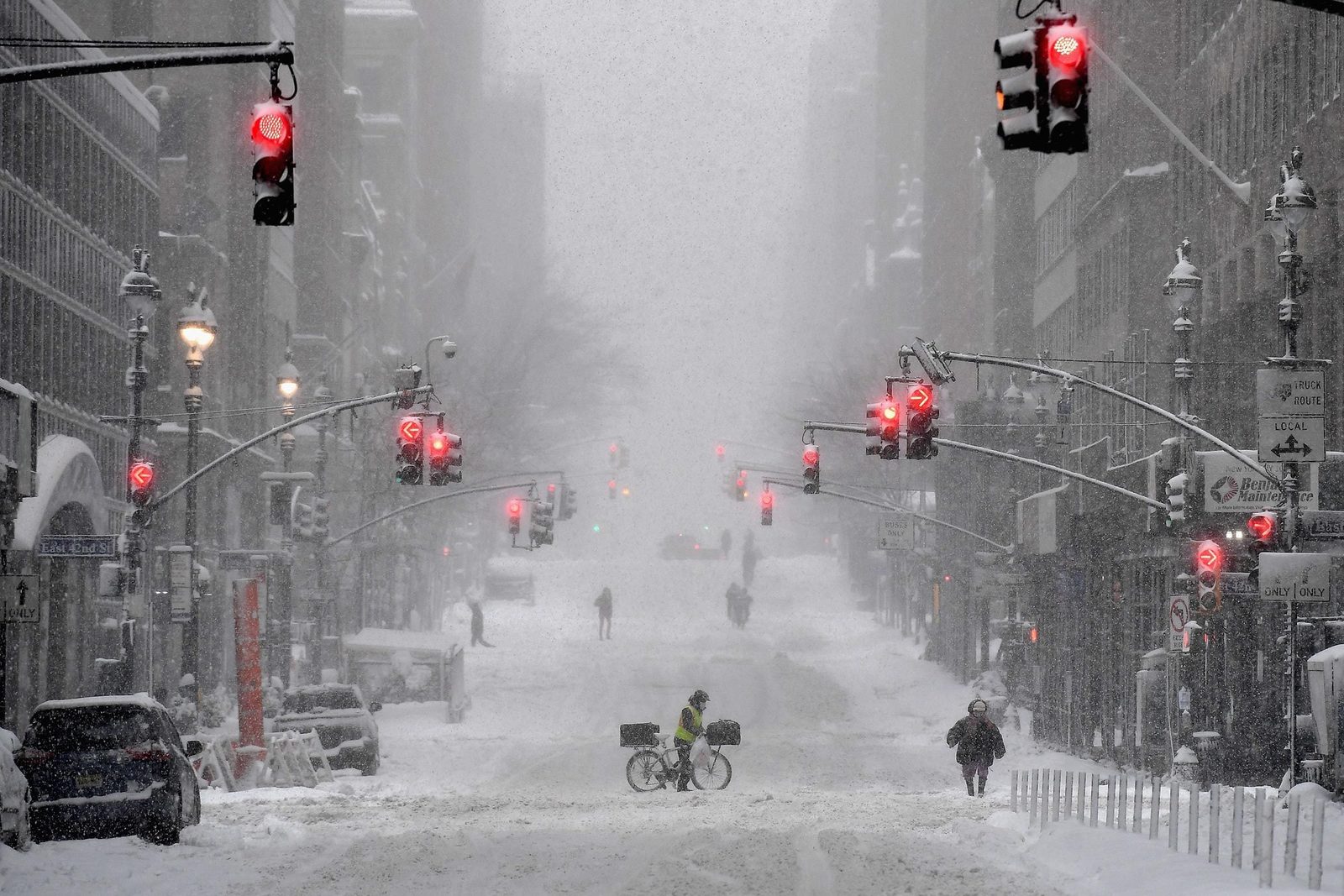 A cyclist stands in snow-covered streets, surrounded by red traffic lights, as pedestrians navigate the blizzard in a deserted urban environ