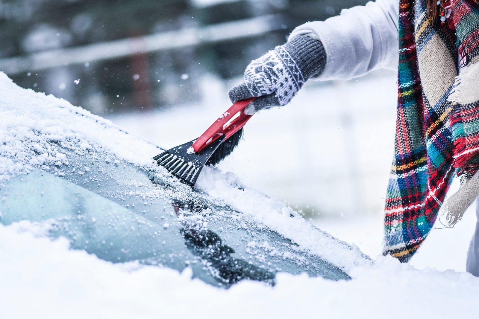 A hand uses a red snow scraper to remove snow from a car windshield in a snowy environment, while wearing a patterned glove and a colorful s