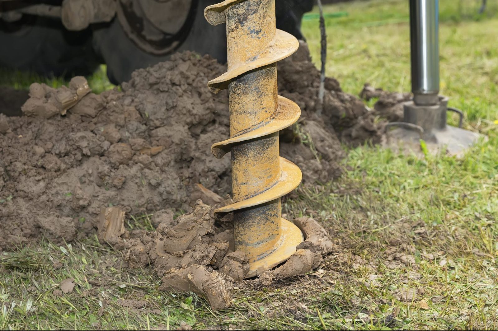 A yellow auger stands in the ground, embedded in dirt, while nearby machinery is partially visible, indicating a drilling operation in a gra