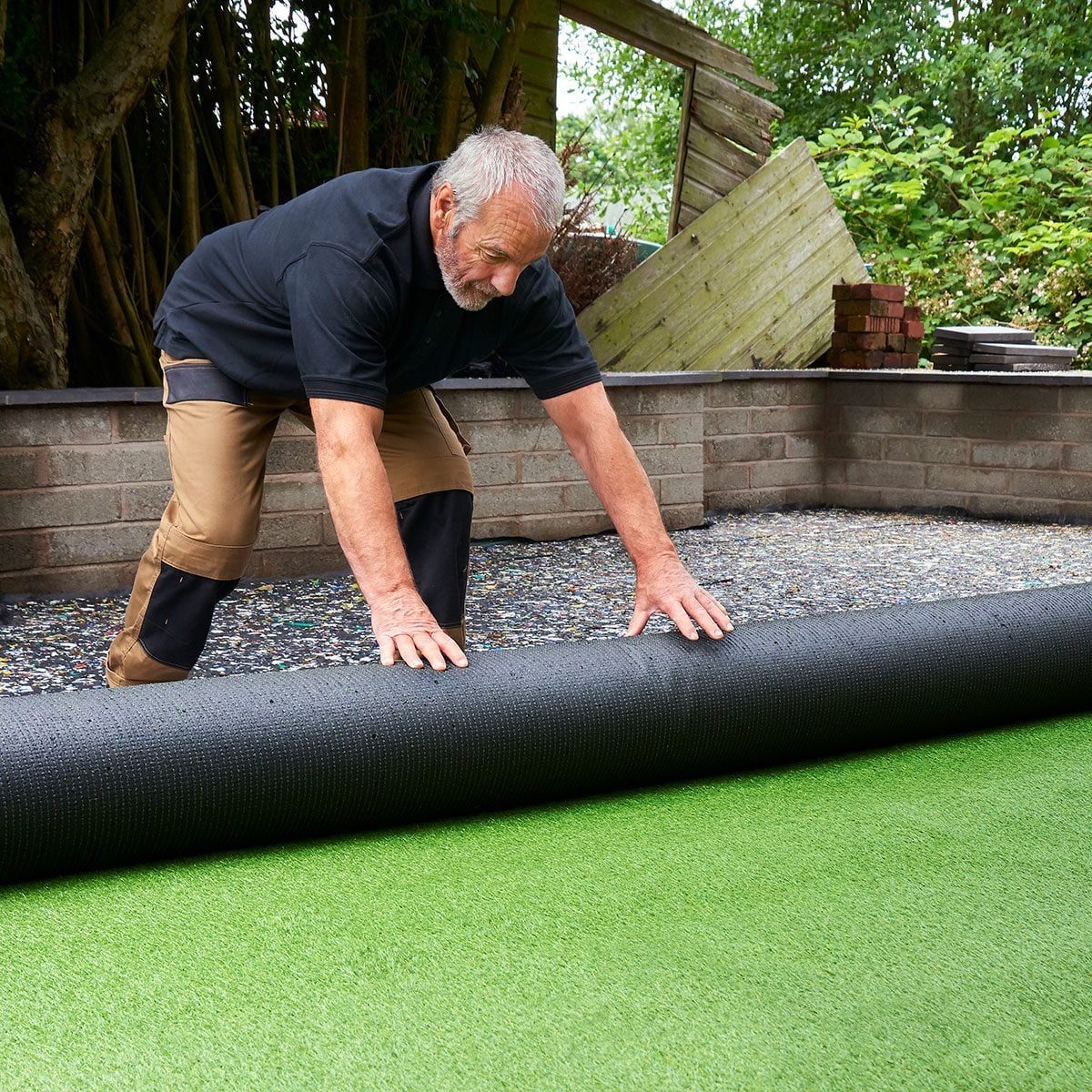 A man rolls out a black mat over green artificial grass in a garden, surrounded by brick walls and trees.