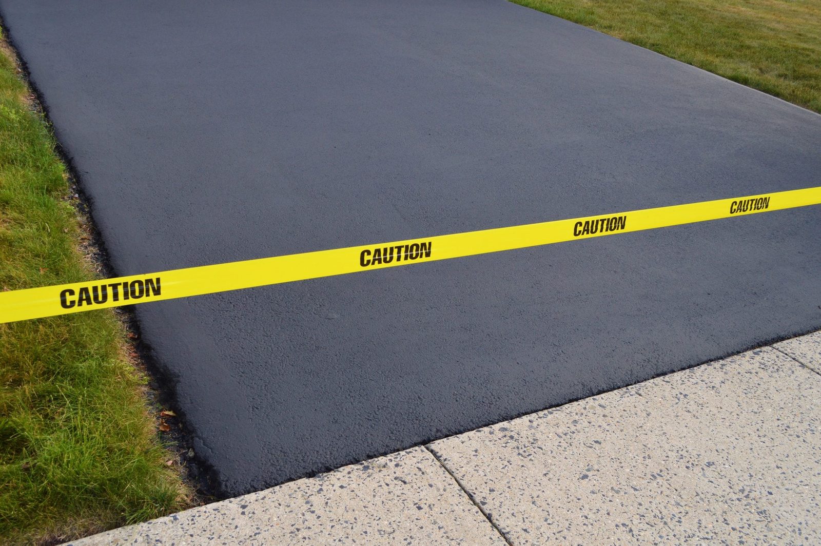 A yellow caution tape lines a fresh, dark asphalt surface, which is bordered by green grass and adjacent to a light-colored concrete path.