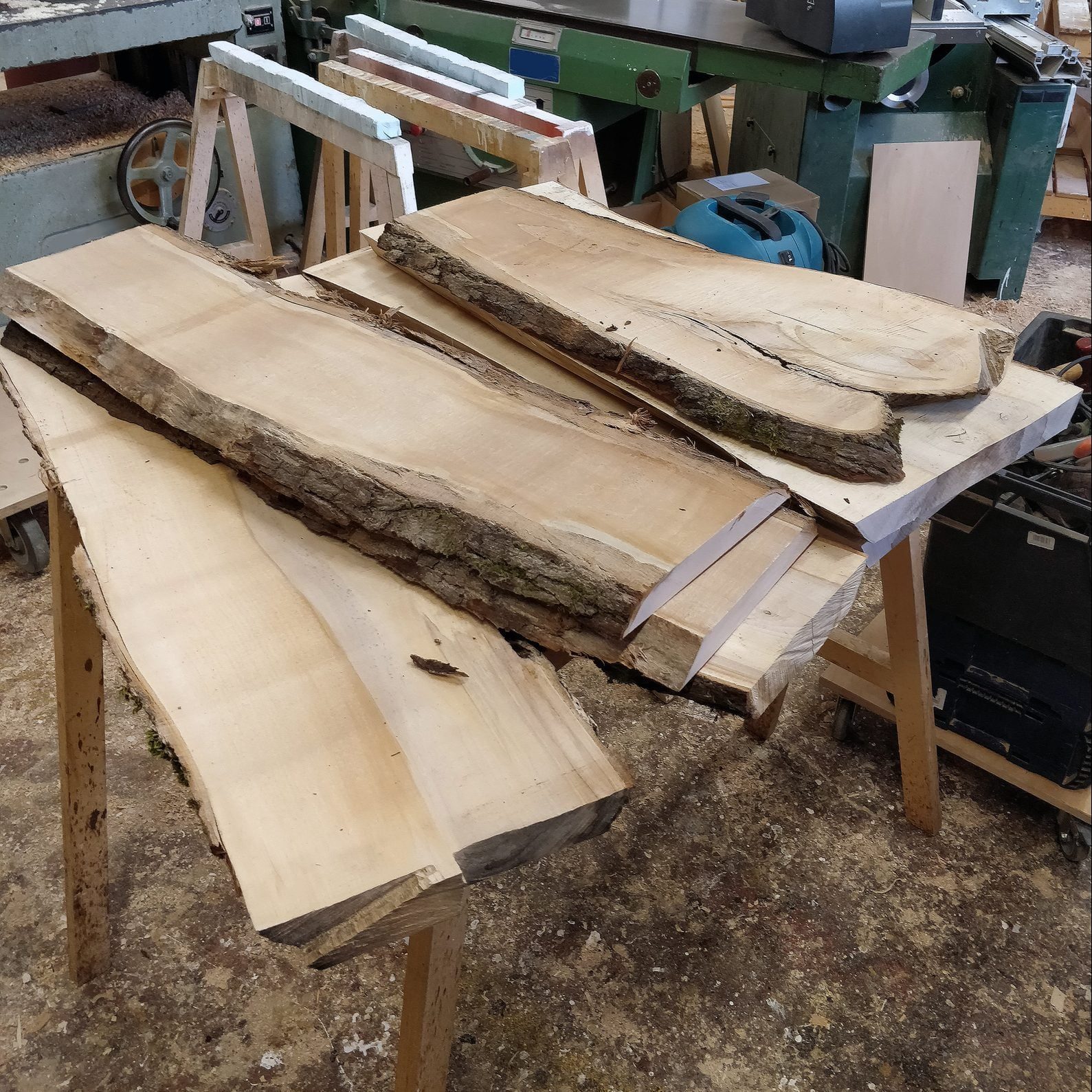 Wooden planks, varying in size, rest on a table in a workshop, indicating recent cutting or preparation. Surrounding tools and equipment sug