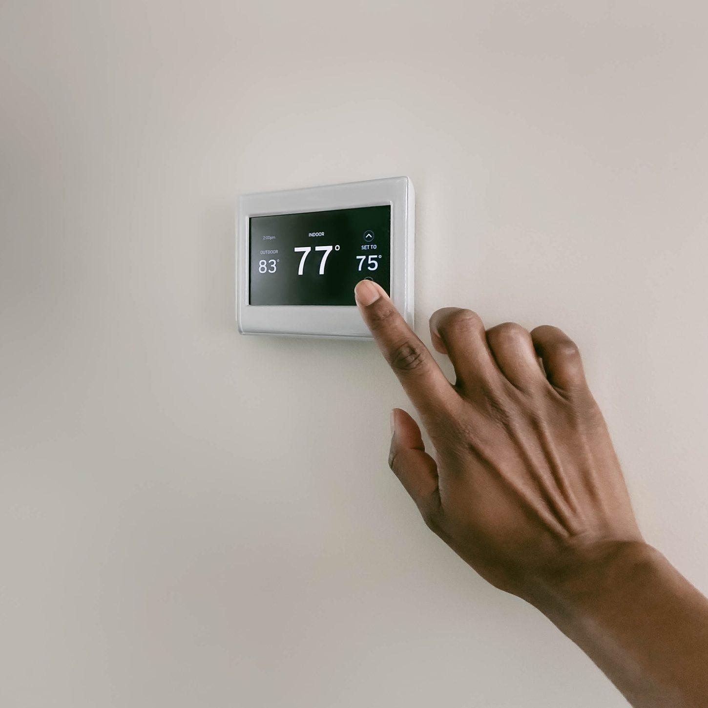 A hand adjusts a thermostat on a wall displaying indoor and outdoor temperatures, with the indoor set to 75°F and currently at 77°F.