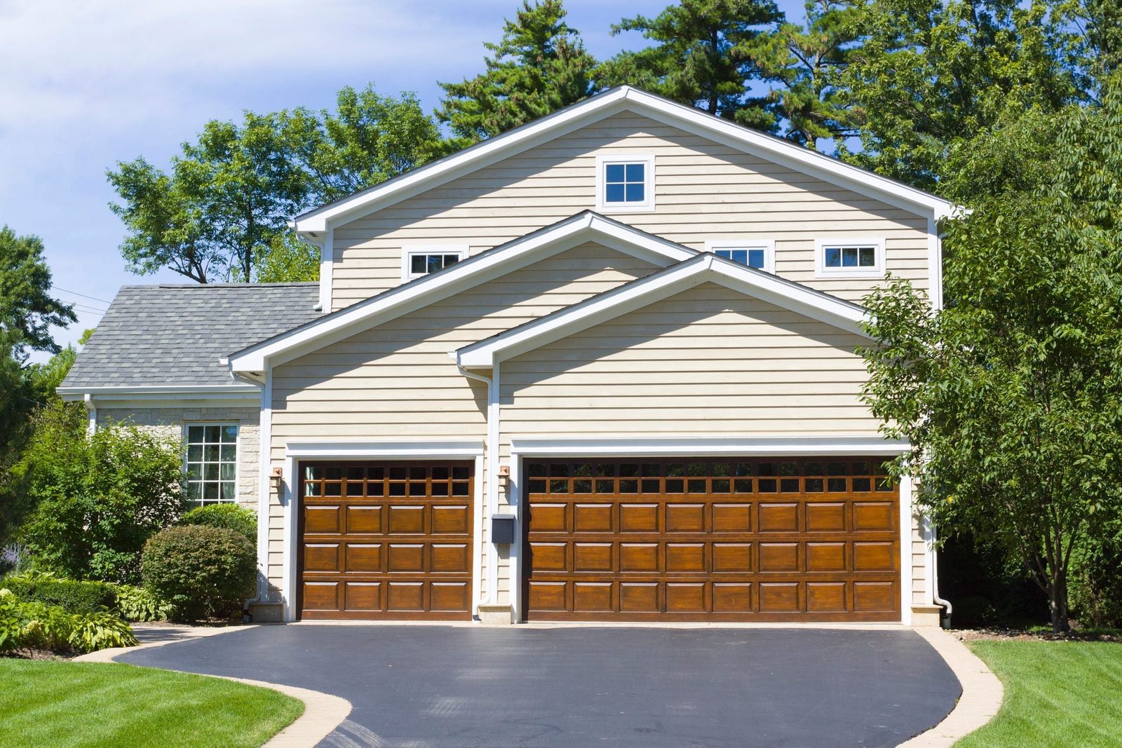 A two-car garage with wooden doors stands at the front of a light-colored house, surrounded by green trees and a well-manicured lawn.
