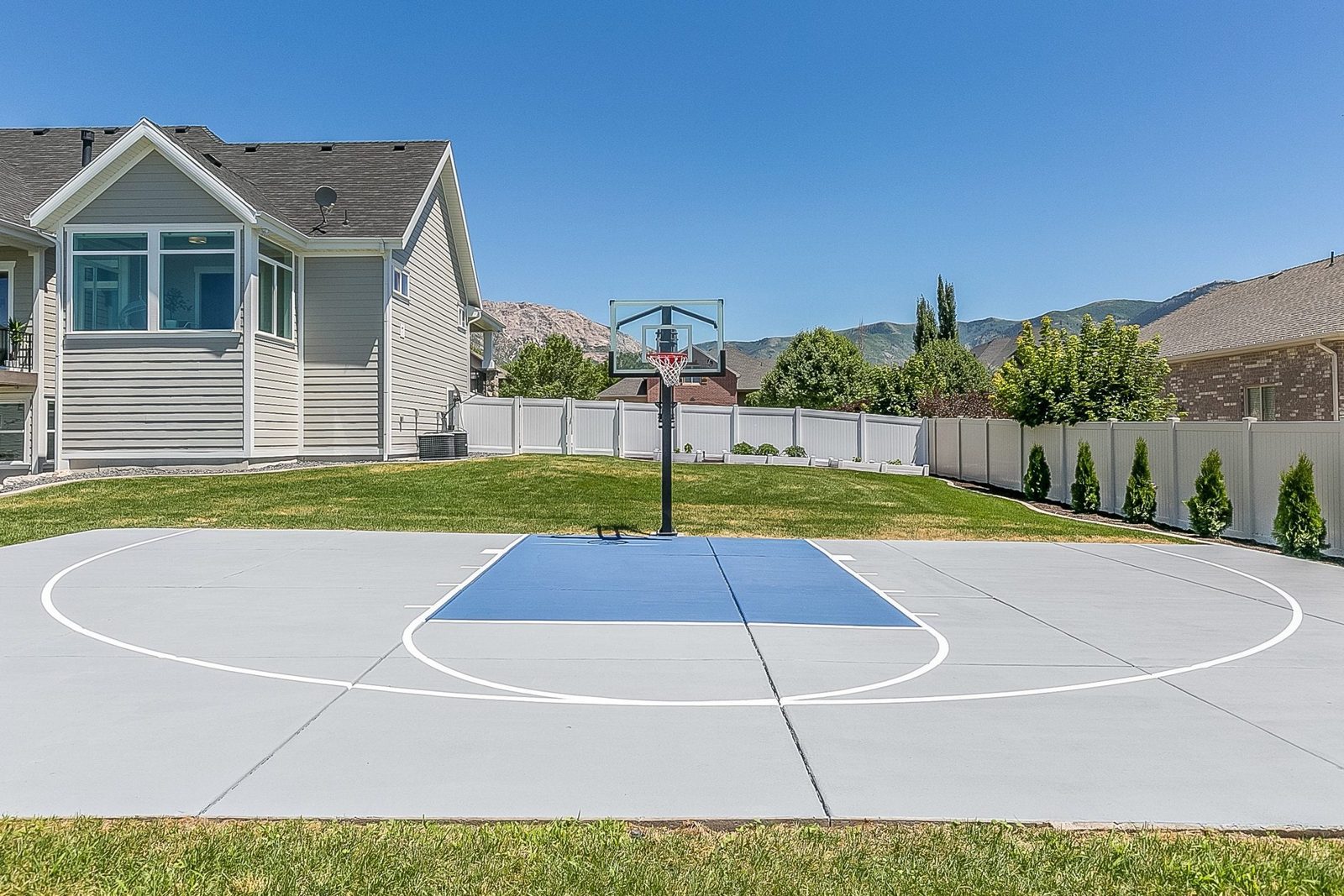 A basketball hoop stands on a blue court in a backyard, surrounded by lush grass and trees, with houses and mountains in the background unde