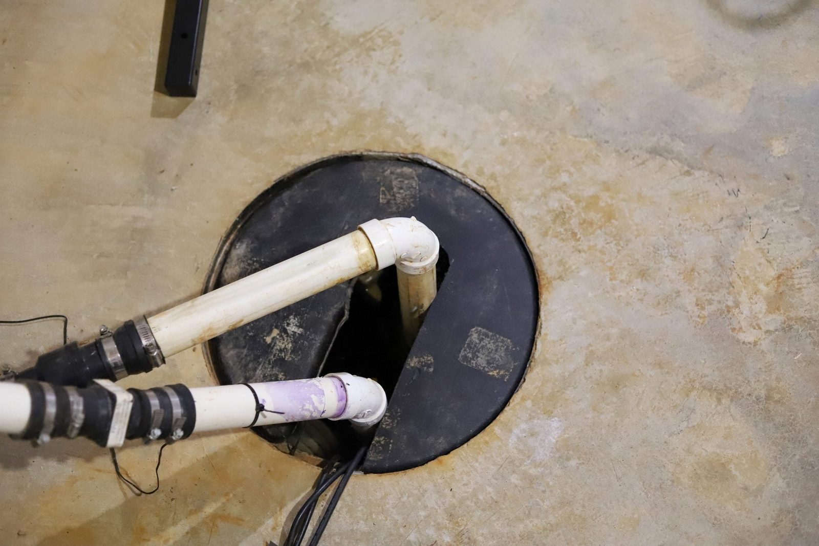 Two PVC pipes connected to a black circular opening extend into a concrete floor, showing worn surfaces and some dirt.