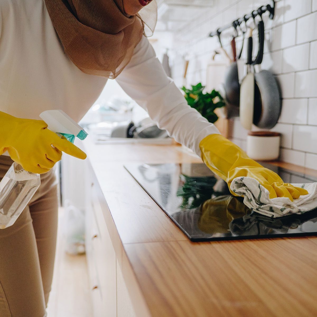 A person in yellow gloves sprays cleaner and wipes a stovetop with a cloth in a bright kitchen, featuring wooden surfaces and hanging utensi