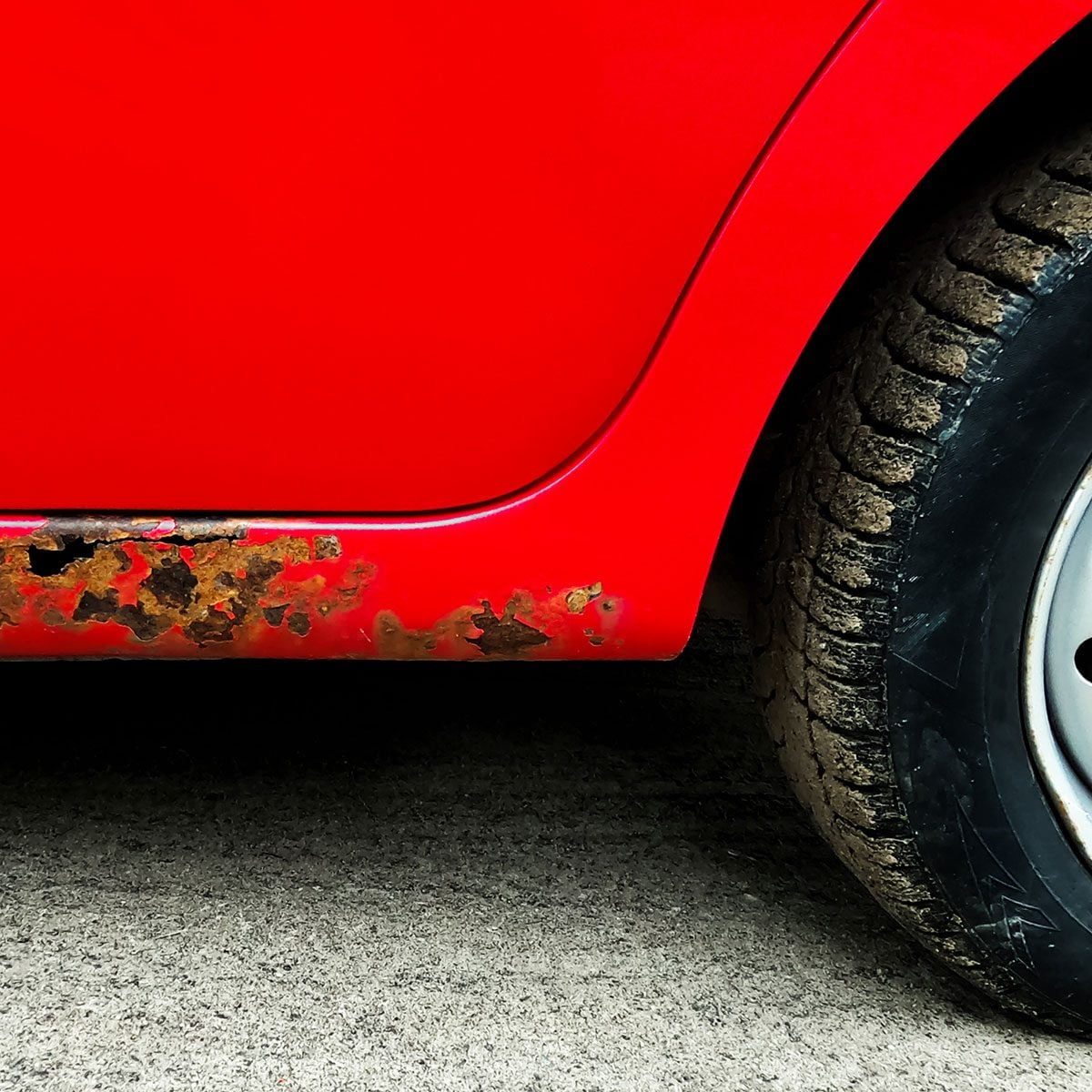 A red car with rust spots along the lower body is parked on a rough, gray surface, and its tire shows signs of wear and mud.