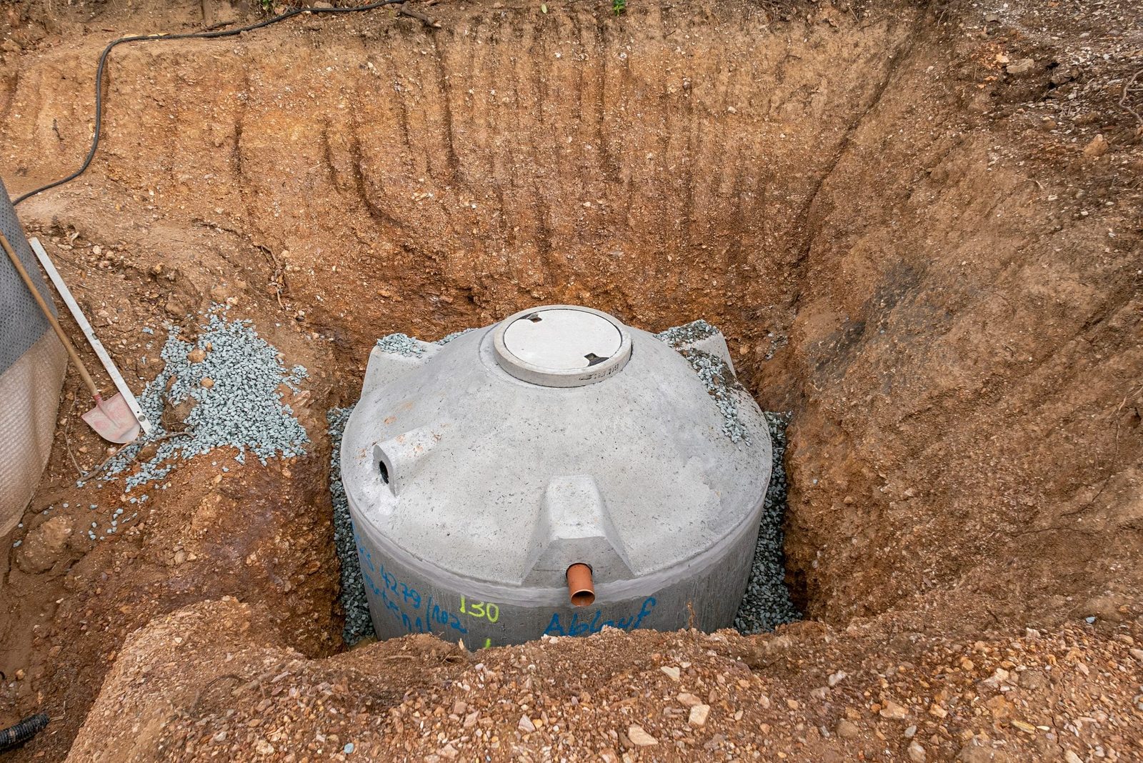 A concrete tank sits at the bottom of a shallow excavation, surrounded by gravel and earth, indicating preparation for installation or maint
