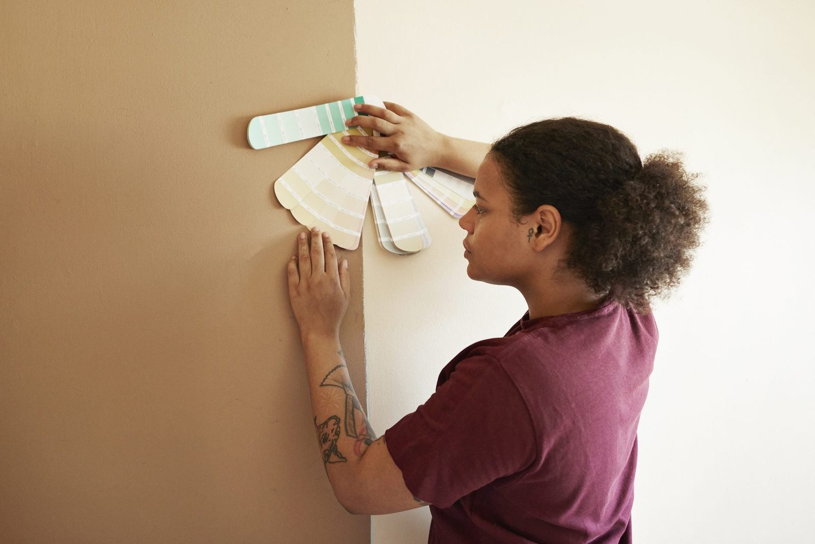 A person holds paint samples against a wall, evaluating colors. The setting features a smooth, light-colored wall next to a corner.