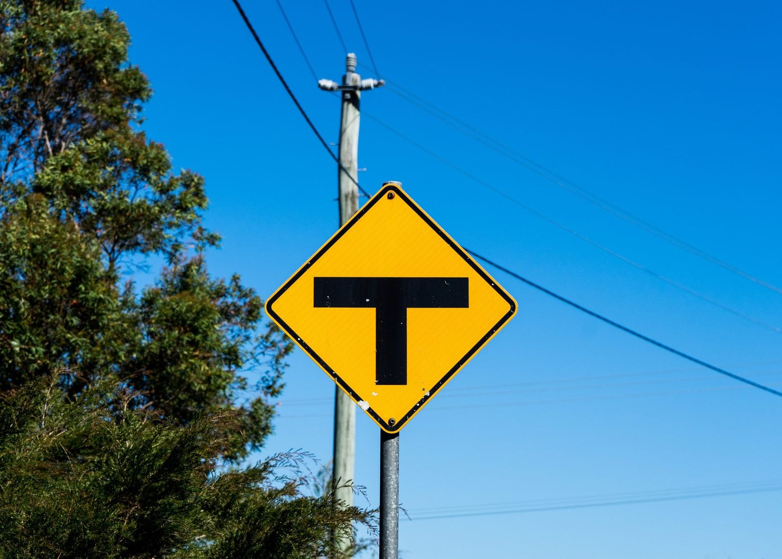 Photo of a black and yellow T intersection road sign with power lines and blue sky in the background