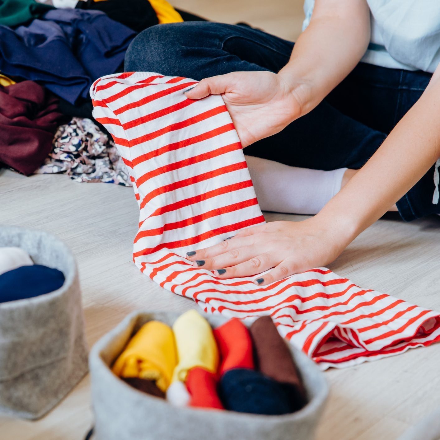 A person folds a red-and-white striped shirt while sitting on the floor, surrounded by neatly organized clothing in baskets.