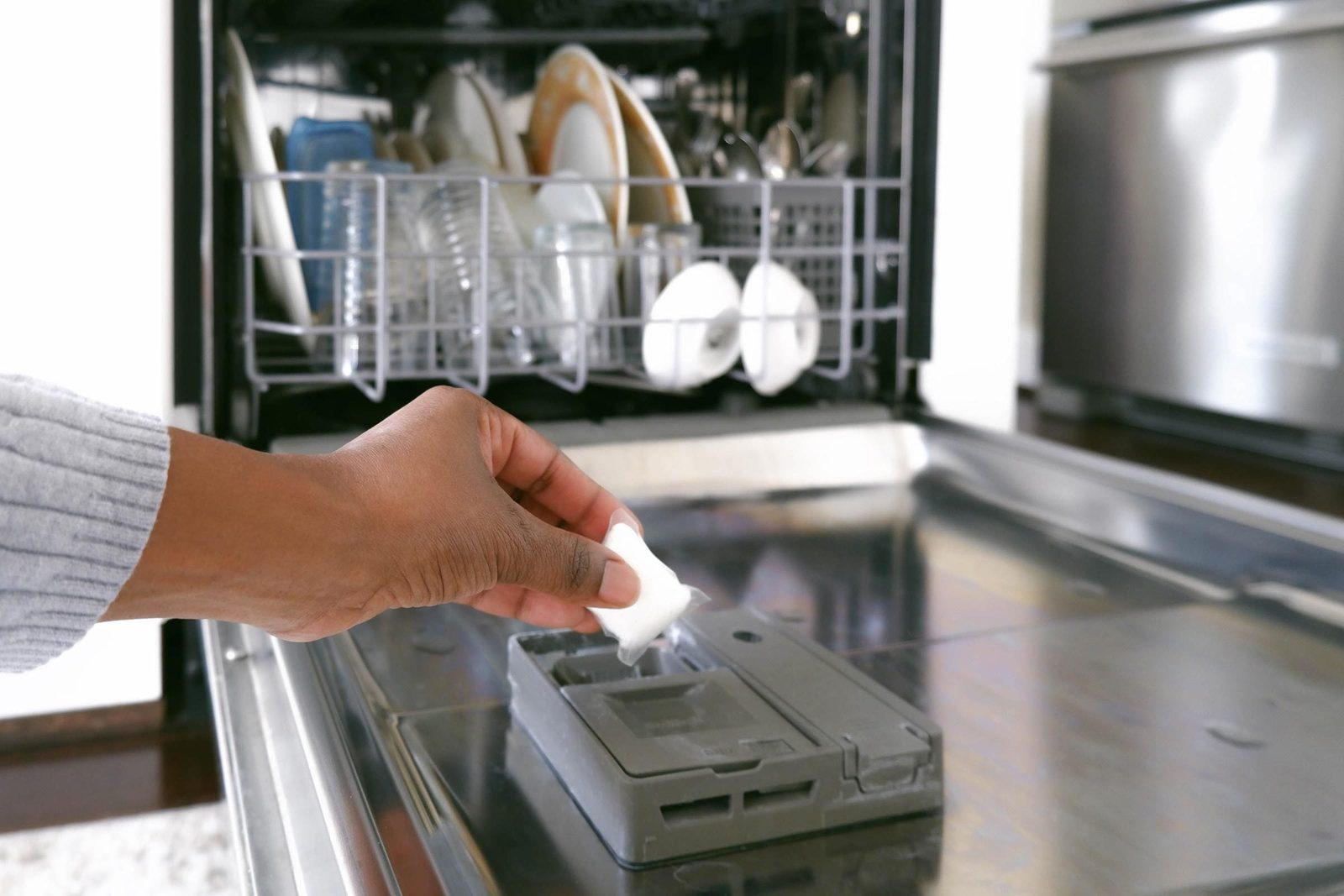 A hand adds a dishwasher pod into the detergent compartment of an open dishwasher, which contains clean dishes and glassware.