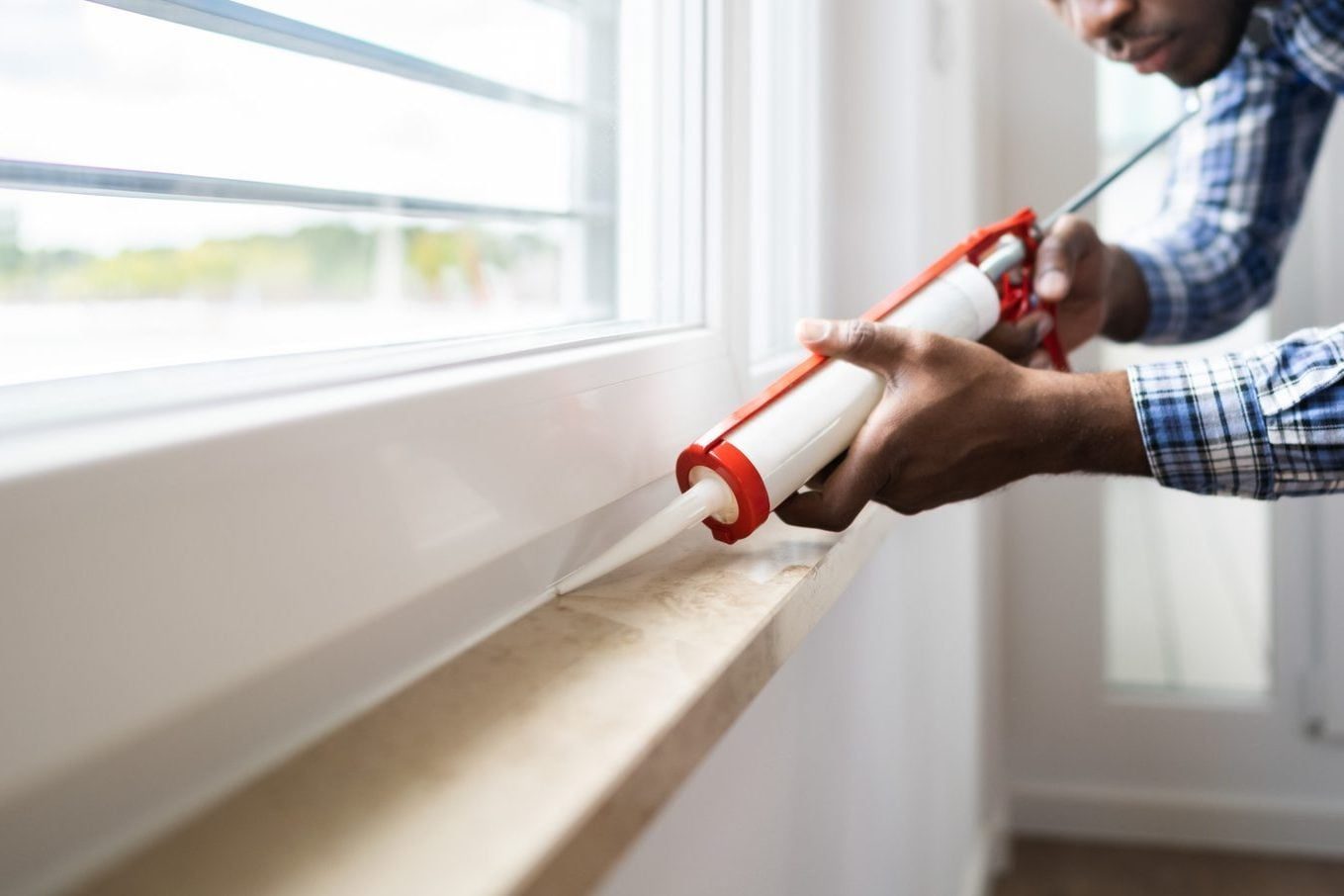 A person applies caulk from a tube along a window frame in a well-lit indoor space, focused on sealing the gap for insulation.